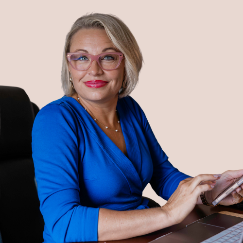 A woman with glasses, blonde hair, wearing a blue dress, sitting at a desk with a smartphone in her hand
