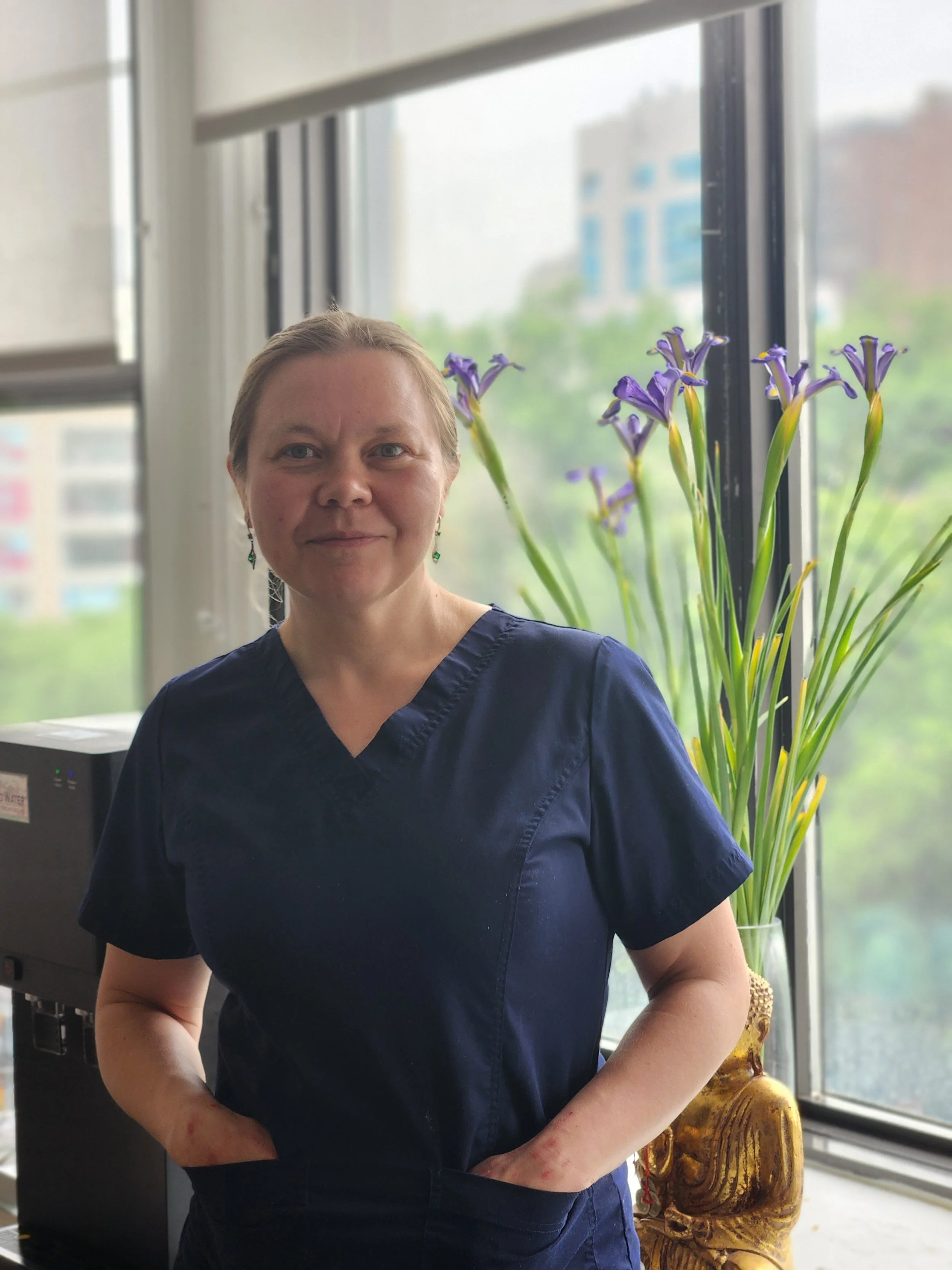A woman wearing a navy scrub top standing next to a large window with a potted purple flower and a wooden carved decoration behind her.