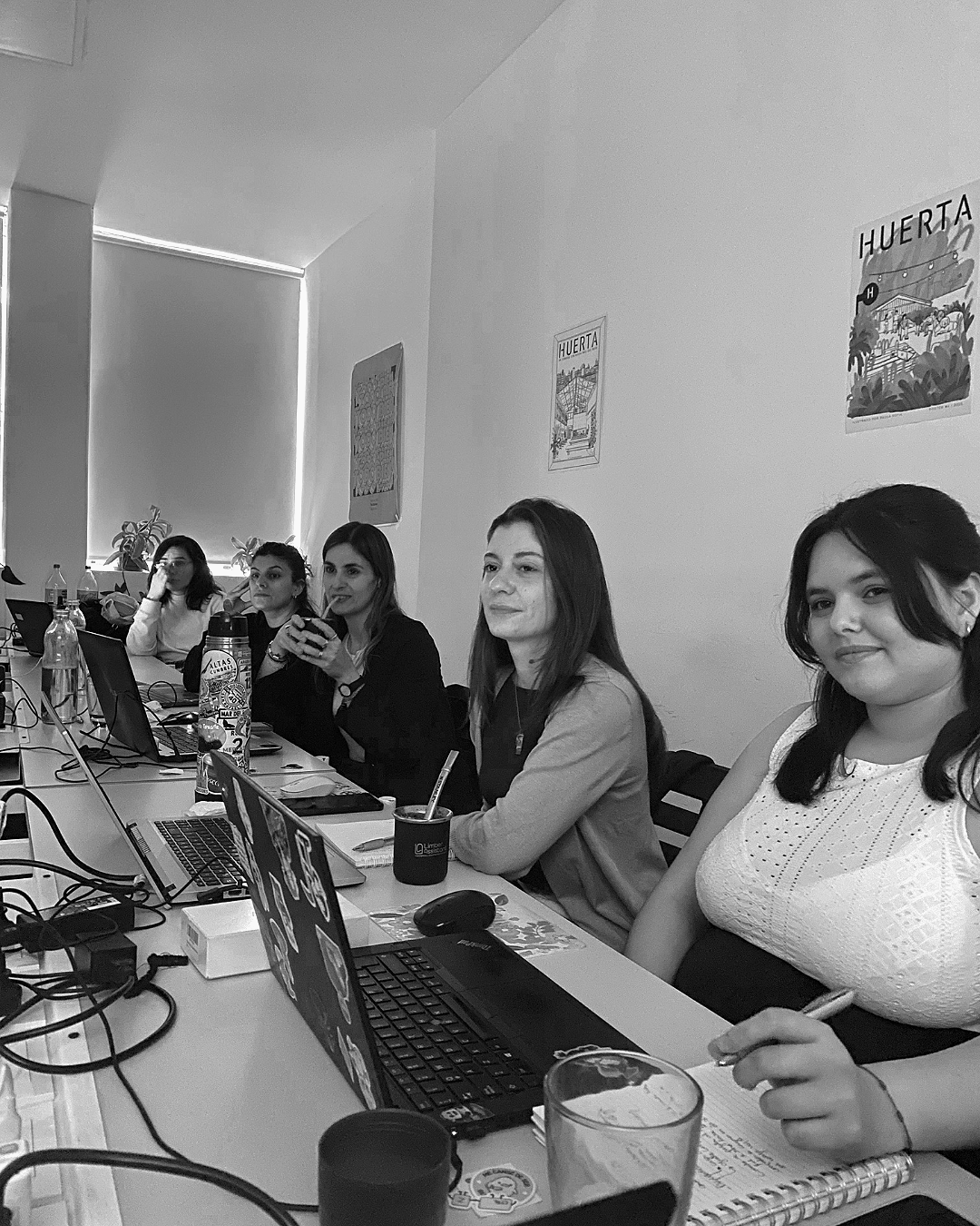 Grupo de cinco mujeres sentadas en una mesa de oficina con laptops, bebidas y material de oficina, en una reunión o trabajo colaborativo, en un espacio cerrado con posters en la pared.