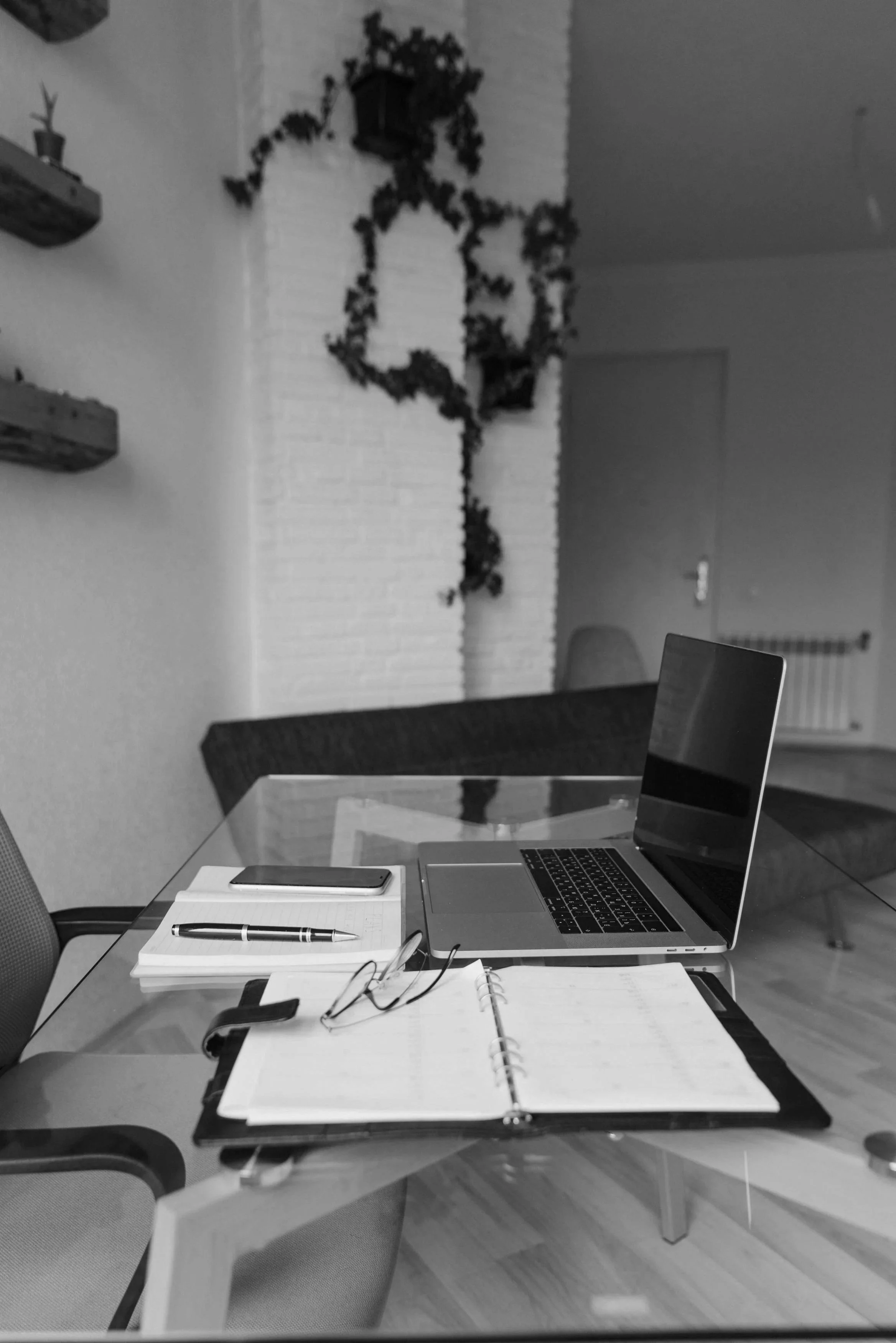 Mesa de oficina con laptop abierta, cuaderno, bolígrafo, teléfono móvil, y gafas. En el fondo, paredes blancas con decoraciones y plantas.