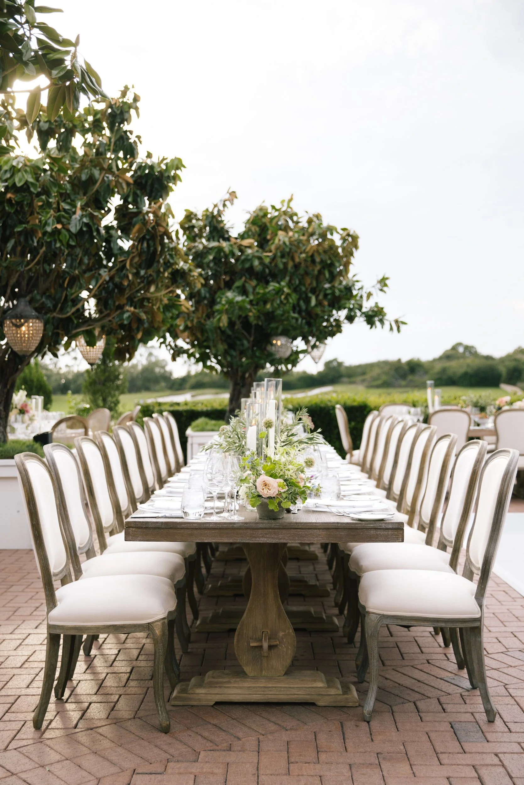 A long rectangular wooden dining table set outdoors during daytime with white cushioned chairs on brick paving. The table has a floral centerpiece with white and blush pink flowers and candles. Overhead, green trees with hanging lanterns provide shad
