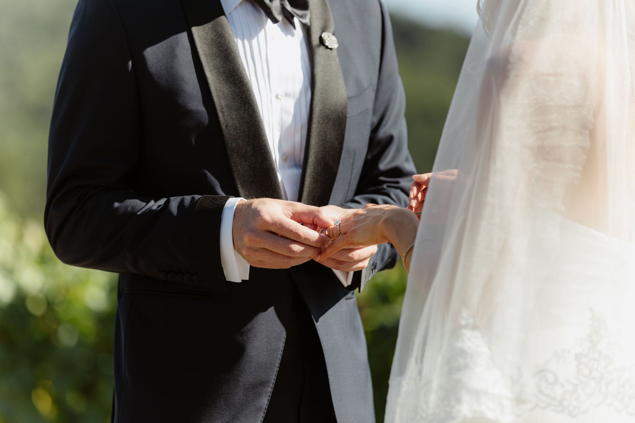 A close-up of a couple exchanging wedding rings outdoors, with focus on their hands and part of their formal attire.