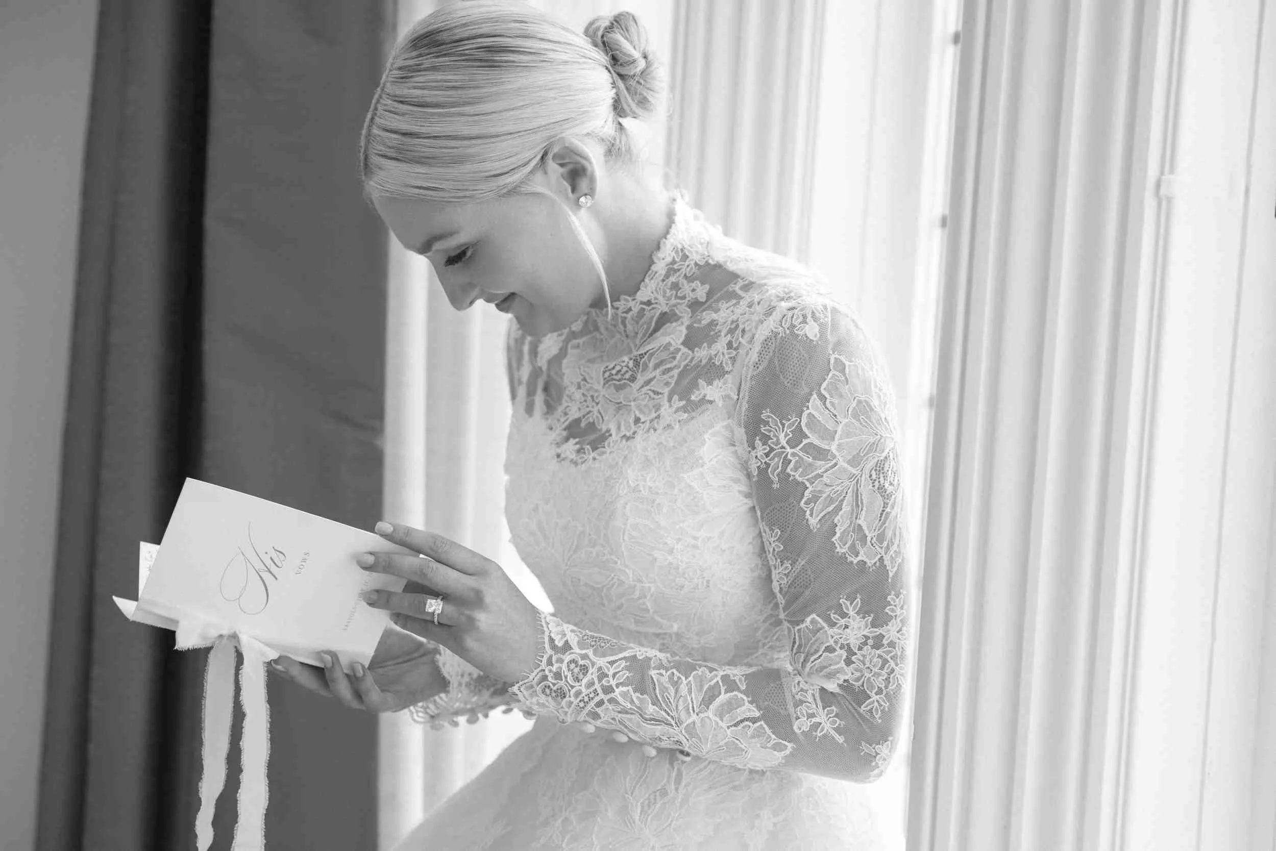 A woman with blonde hair styled in an elegant updo, wearing a lace dress, is reading a card while standing next to a window with curtains.