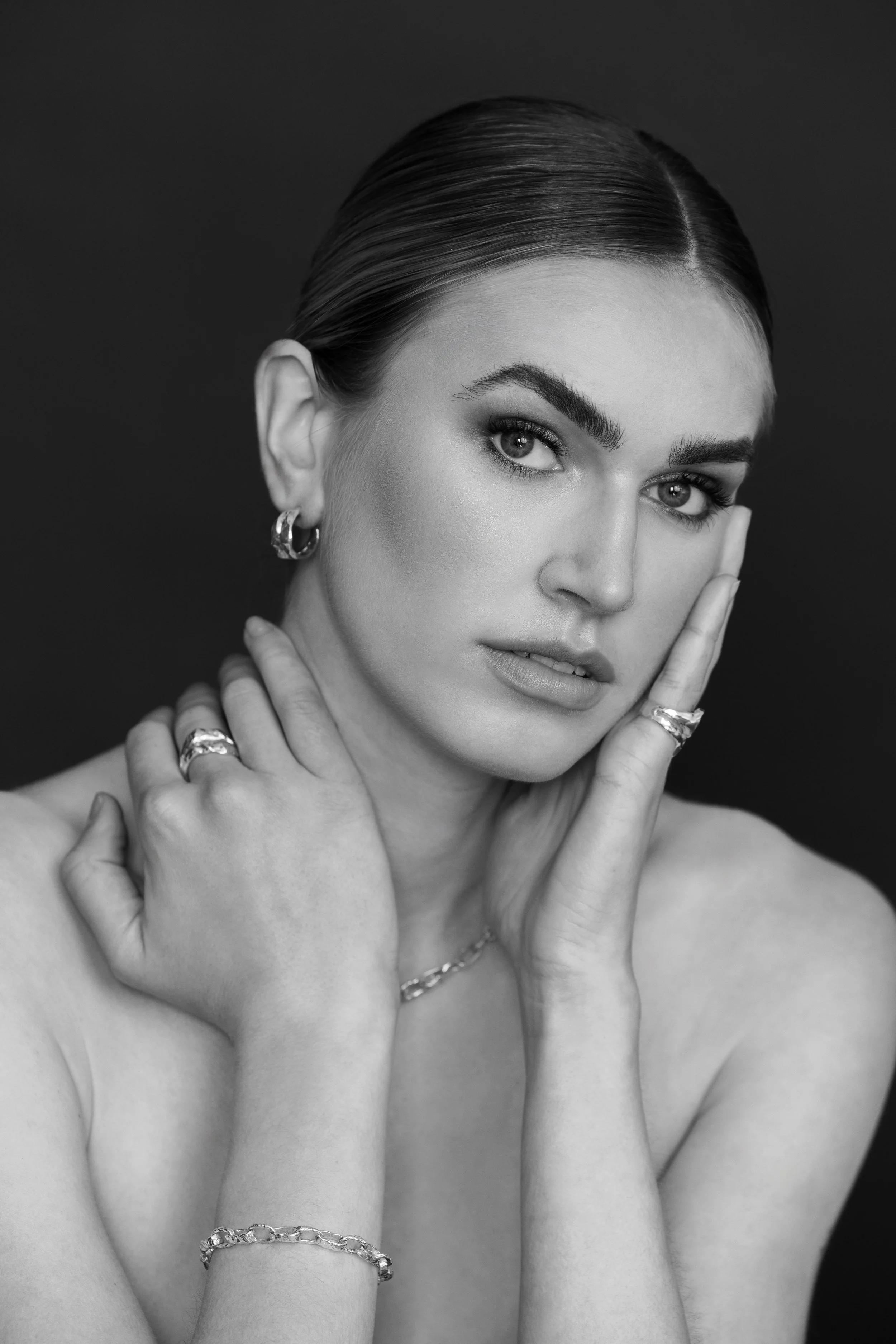Black and white portrait of a woman with slicked-back hair, intense gaze, wearing rings and bracelets, touching her face and shoulder.