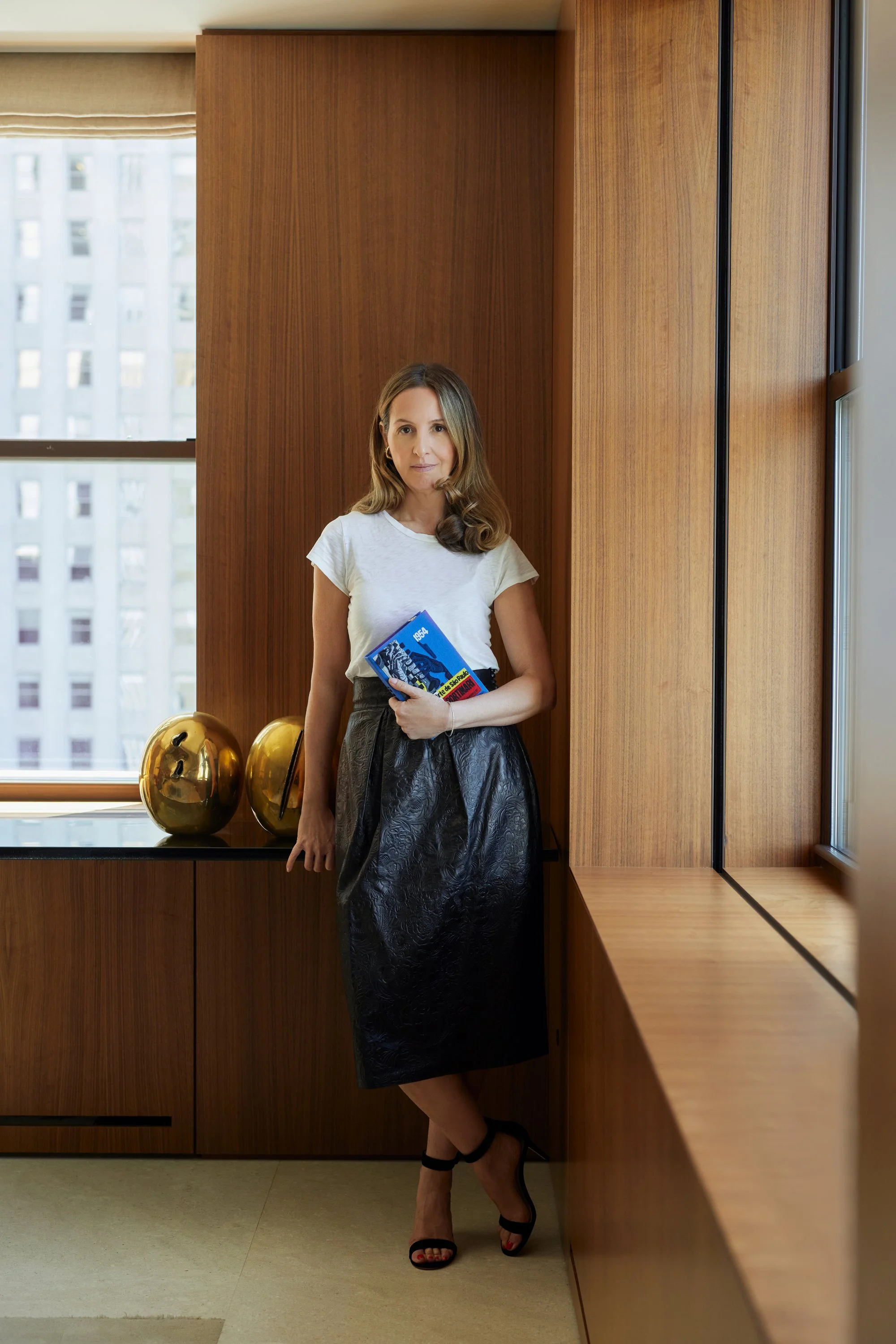 A woman wearing a white t-shirt and a black skirt with heels, standing in a room with wooden walls and large windows, holding a blue book.
