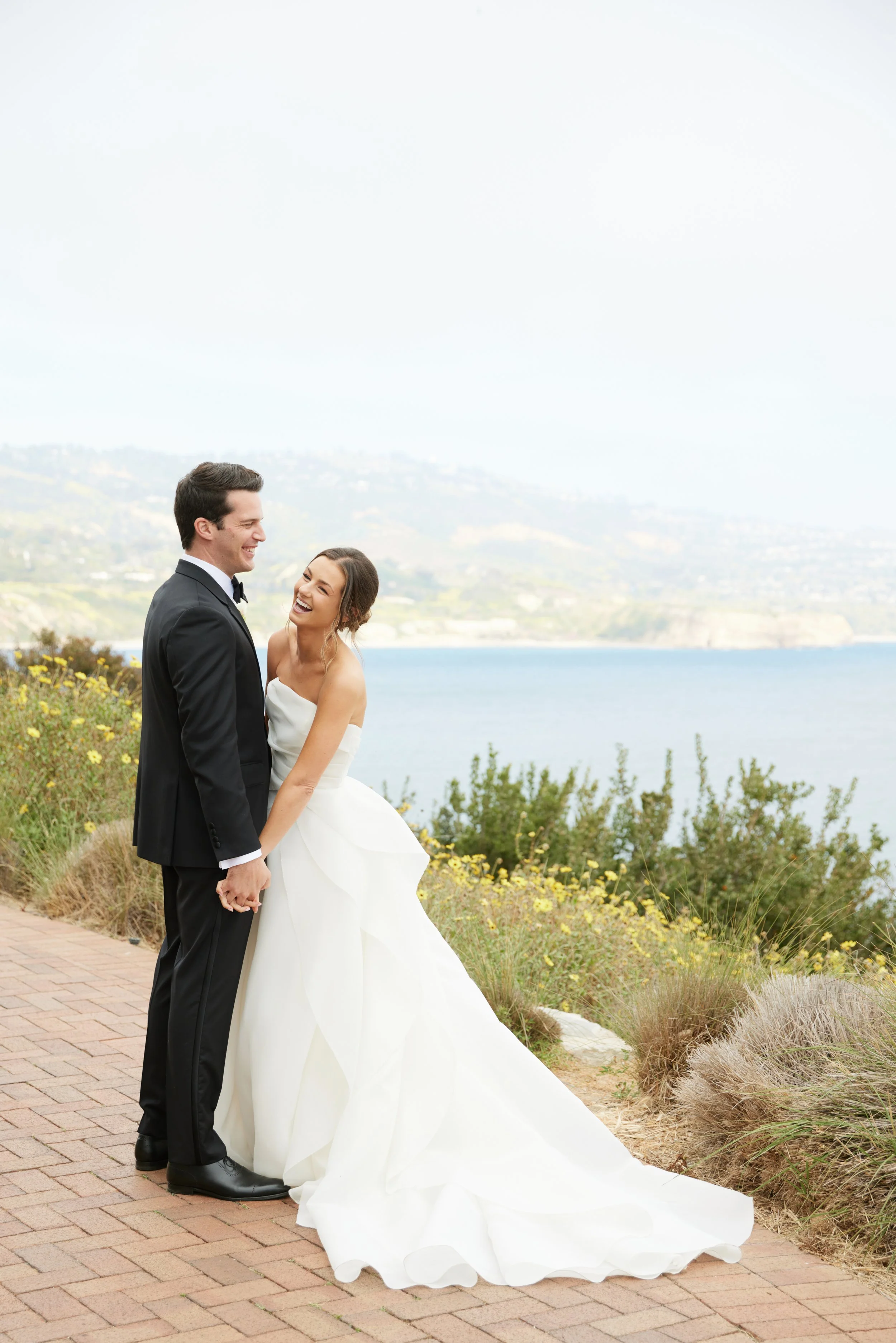 A happy bride and groom sharing a moment outdoors near a lake with a scenic view, smiling and holding hands.