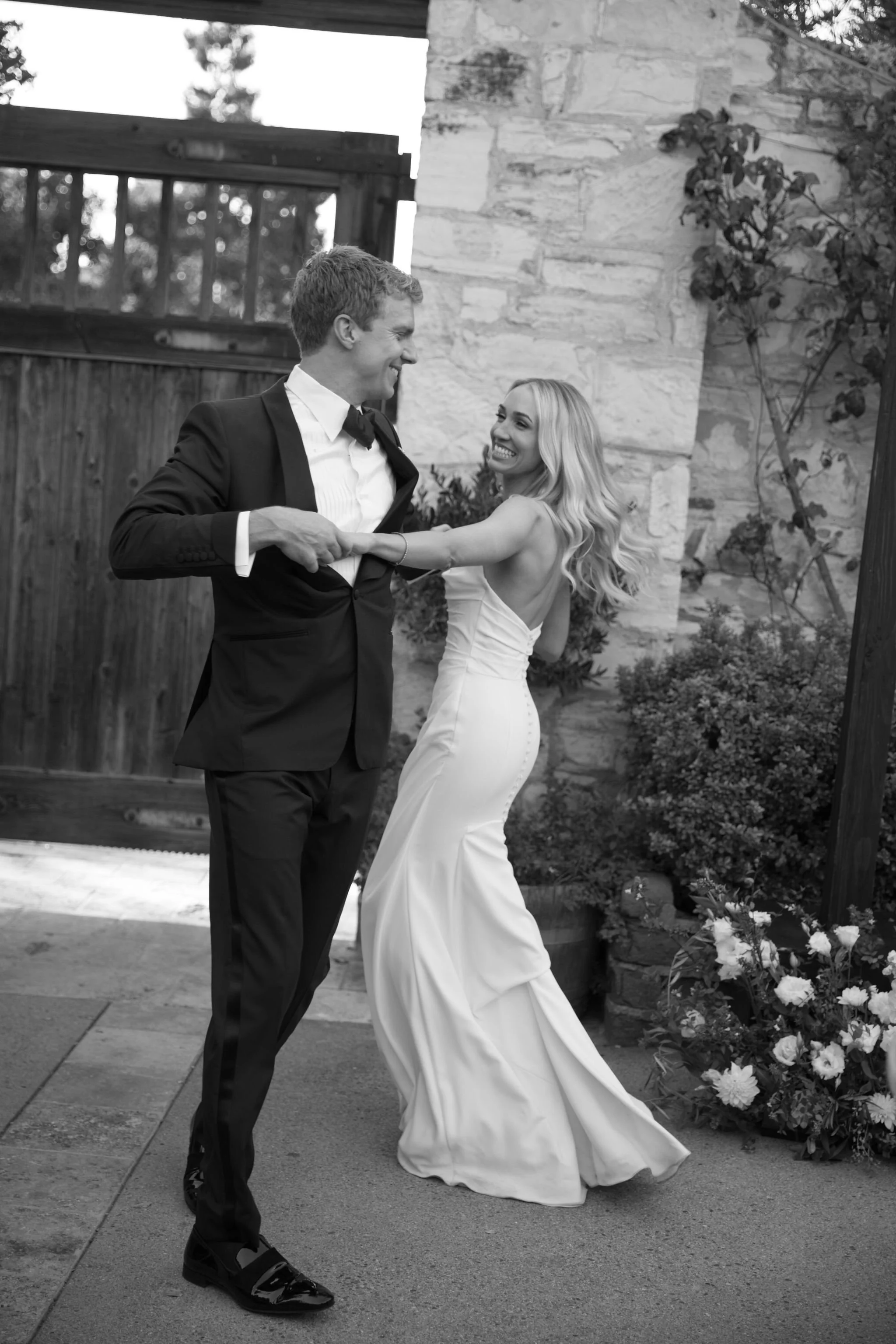 A bride and groom dancing outdoors in front of a stone wall and garden, smiling and enjoying their wedding celebration.