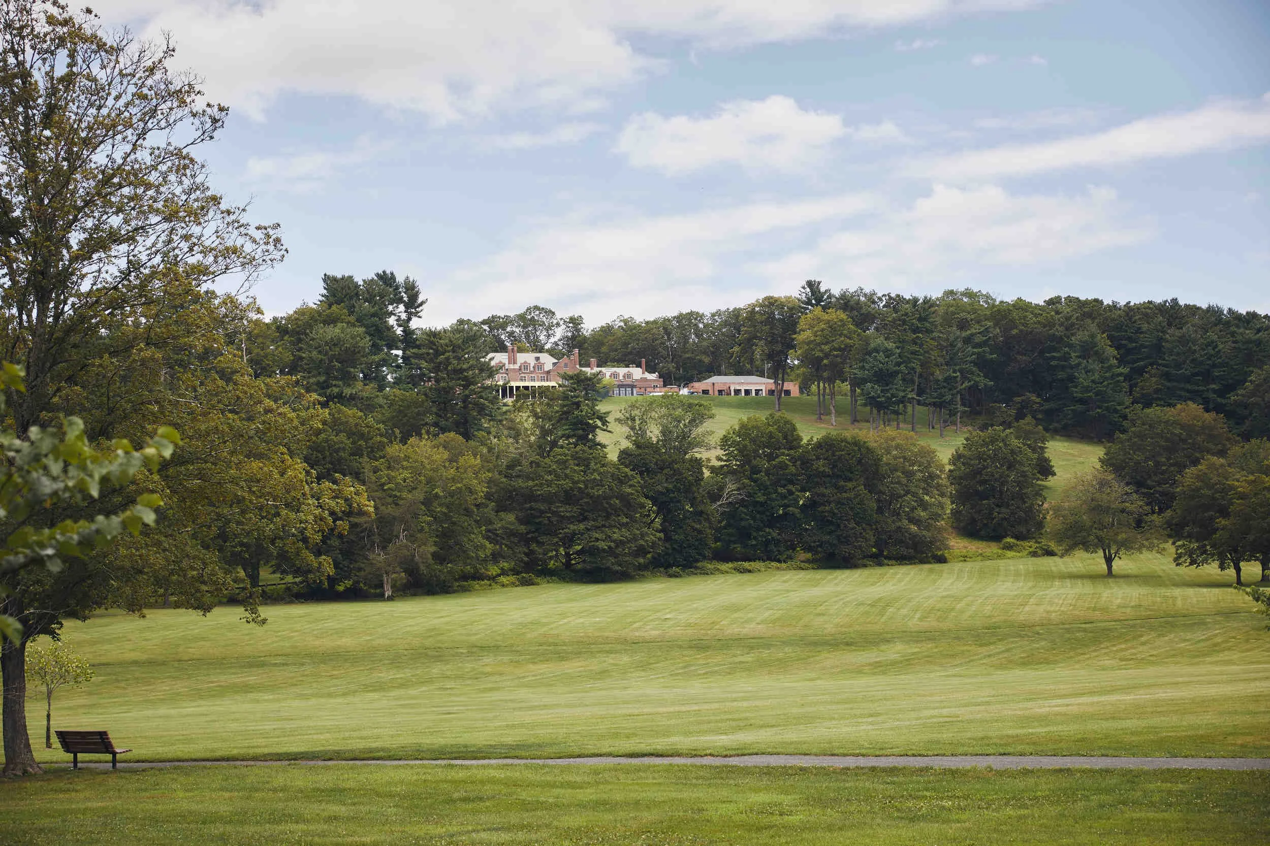 A scenic view of a large house on a hill beyond a lush green golf course or park with trees and a bench in the foreground under a partly cloudy sky.