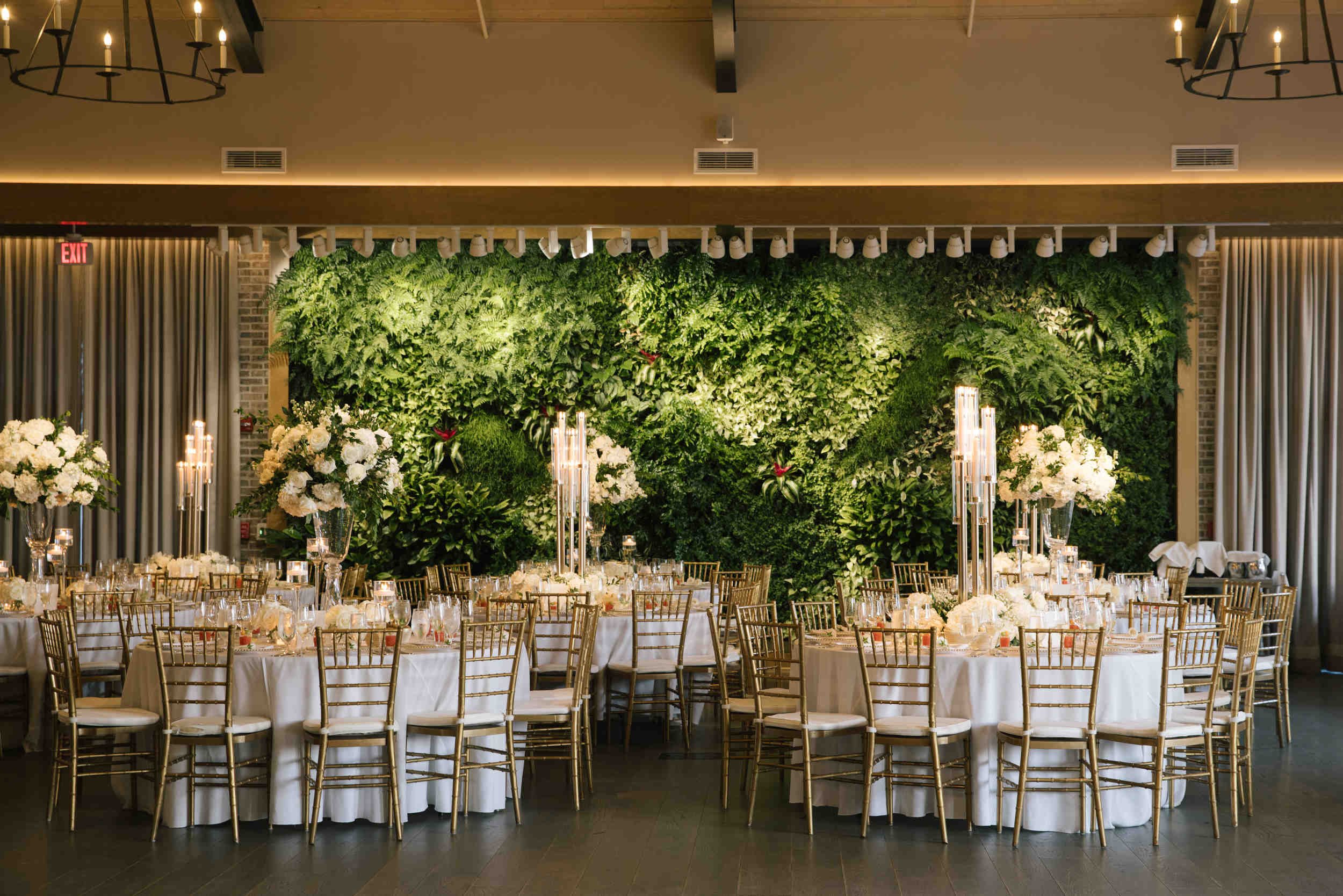 Elegant event space with round tables covered in white tablecloths, decorated with candles and floral centerpieces, set for a formal occasion. A lush green wall with plants is in the background, framed by dark curtains and brick columns. Overhead lig