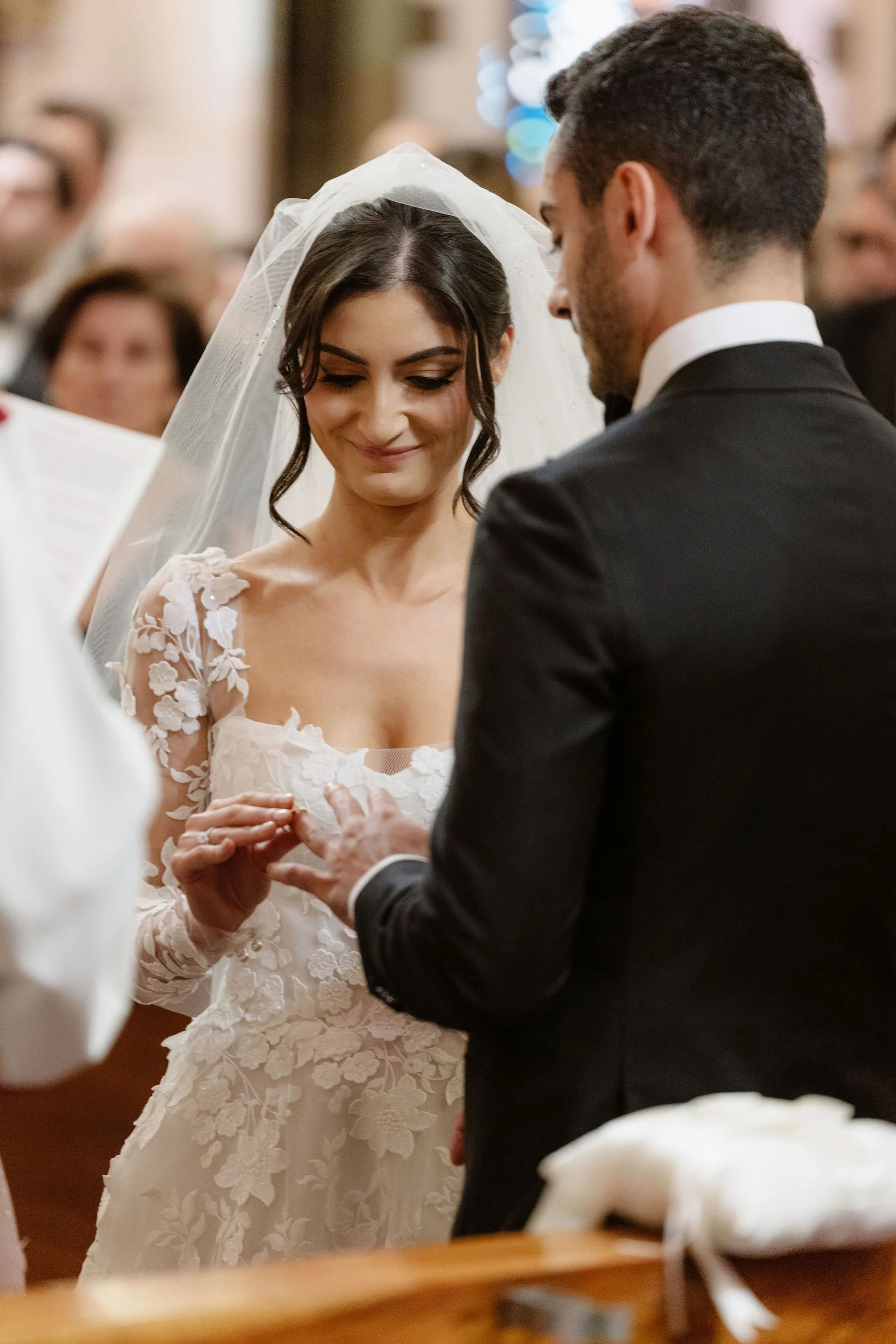 A bride and groom exchanging rings during a wedding ceremony in a church. The bride is wearing a white lace wedding dress with floral details, a veil, and has dark hair. The groom is wearing a black suit with a white shirt. Several guests are blurred