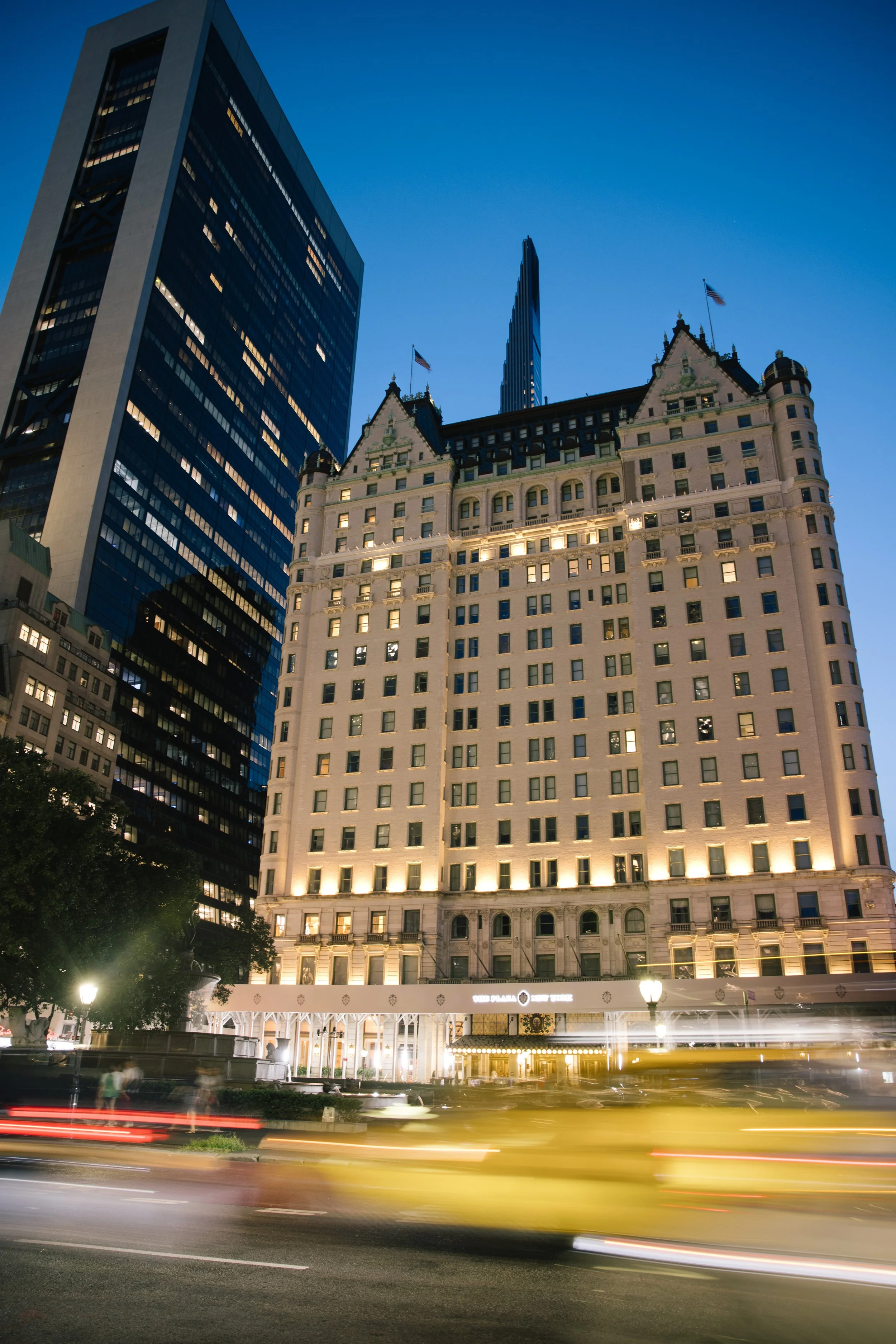 Nighttime cityscape with tall historic hotel building in the foreground, modern glass skyscraper to the left, and a blue sky above, with light streaks from moving cars on the street.
