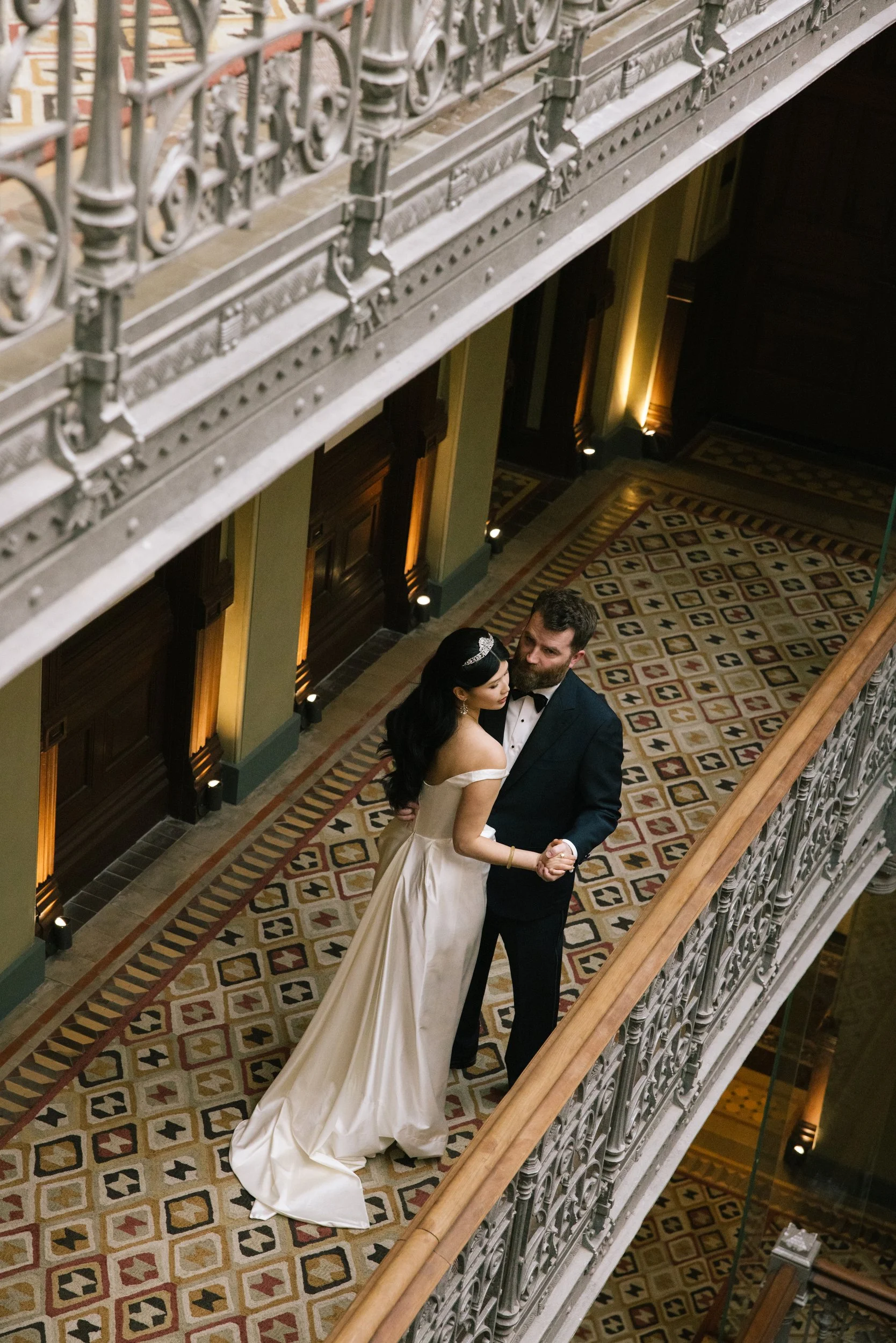 A couple in wedding attire sharing a dance on a patterned carpeted floor in an elegant indoor setting. The woman is wearing a strapless cream-colored gown and the man is wearing a tuxedo. They are holding hands and looking at each other, with decorative lighting on the walls and ornate ironwork balcony railing above.