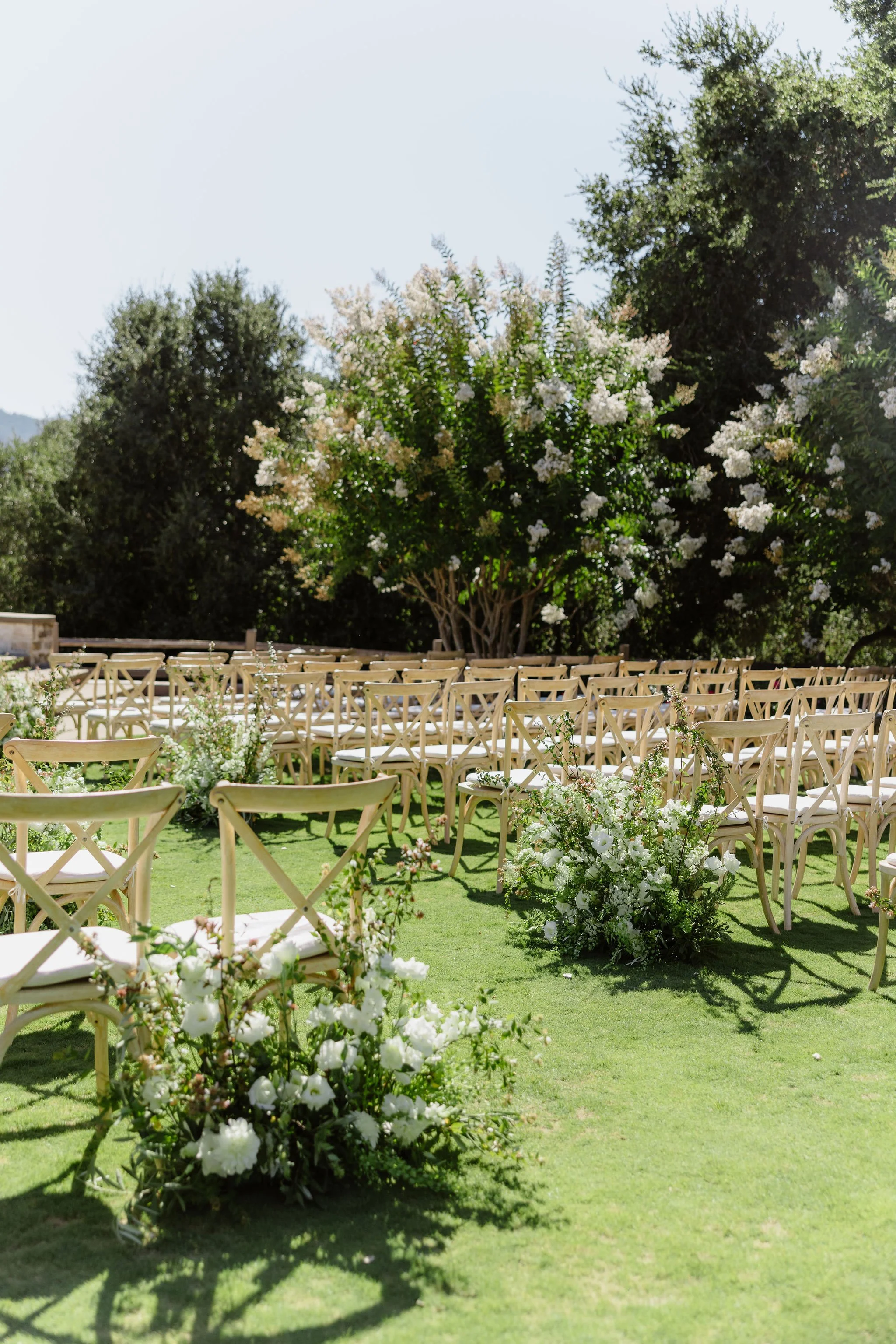 Outdoor wedding ceremony setup with white chairs, floral arrangements, and lush green trees under a clear sky.