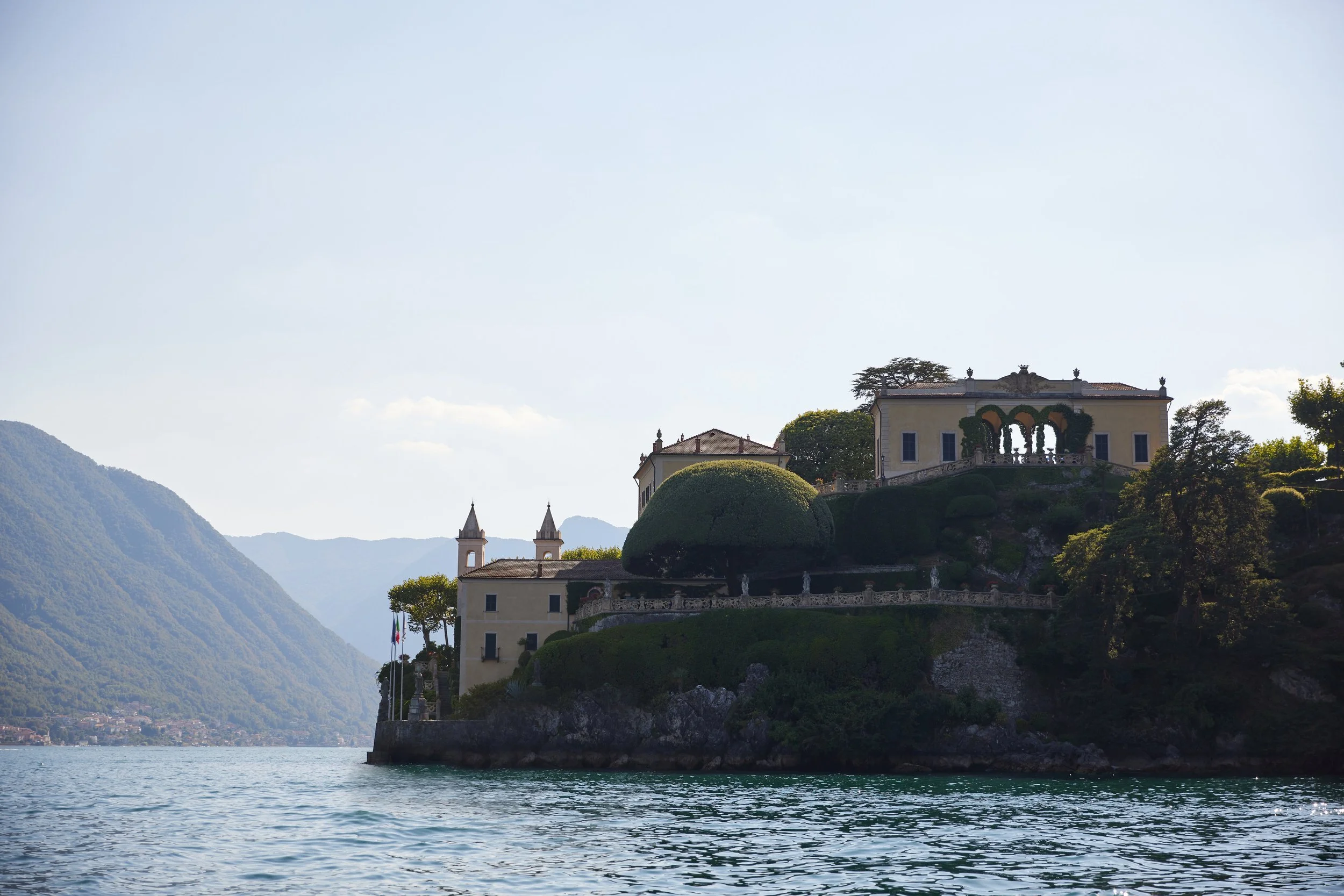 A lakeside view of an elegant mansion built on a hillside with balconies and ornate architecture, surrounded by greenery and overlooking a large lake with mountains in the background.
