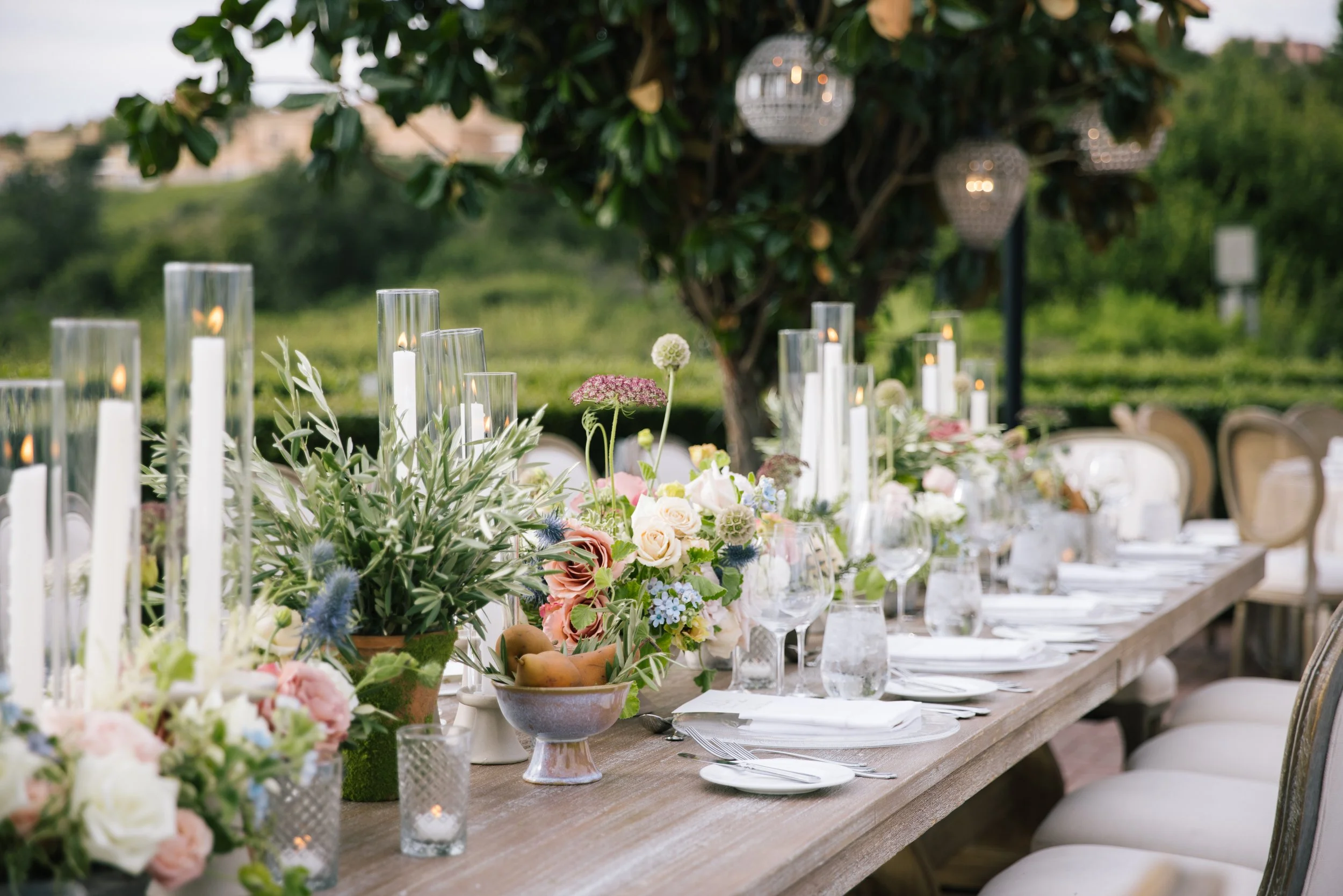 A long wooden table decorated with flowers, candles, and tableware set for an outdoor event, with trees and hanging lanterns in the background.