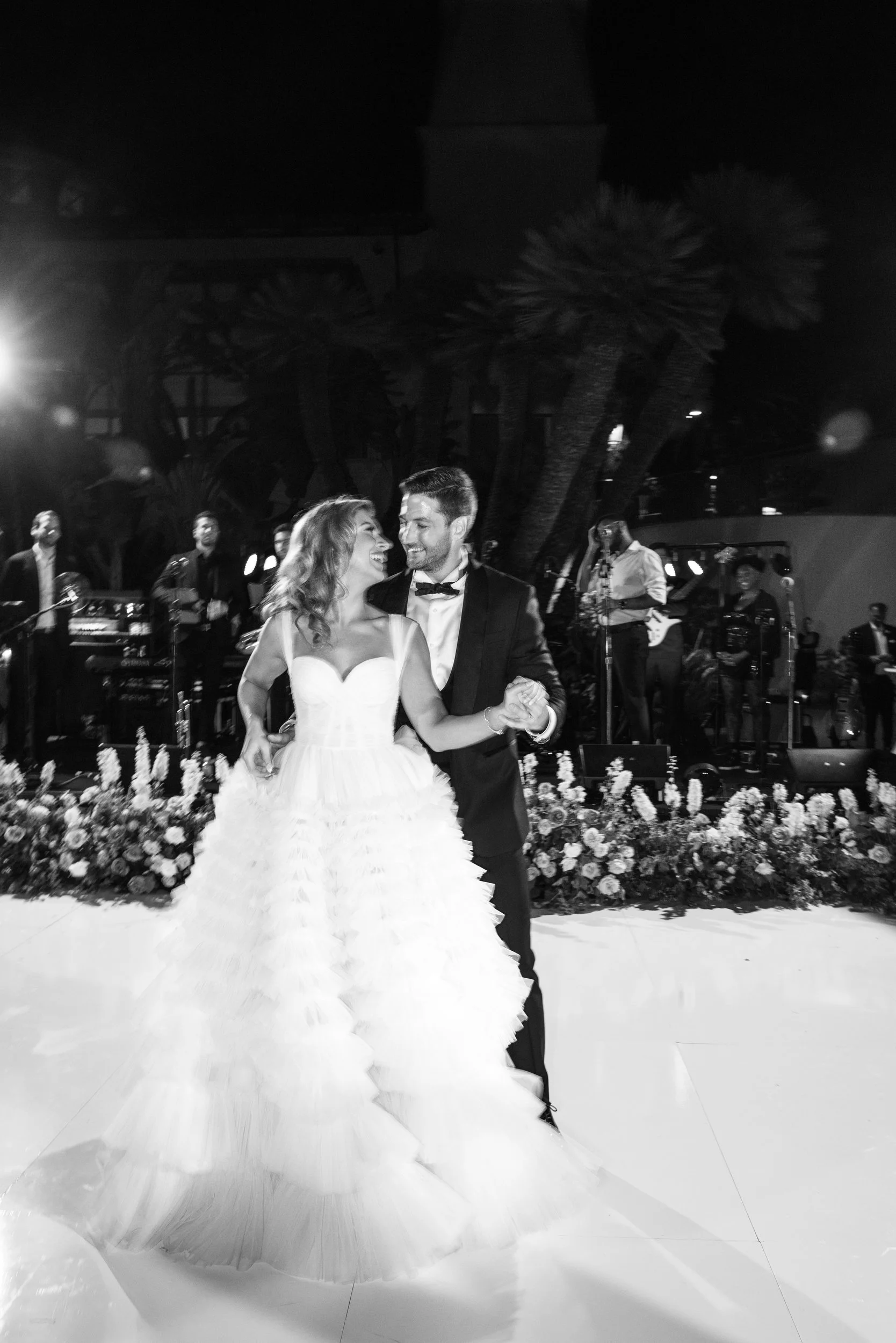 A bride and groom dancing at a wedding reception at night, with a live band performing on stage in the background and floral decorations along the stage edge.
