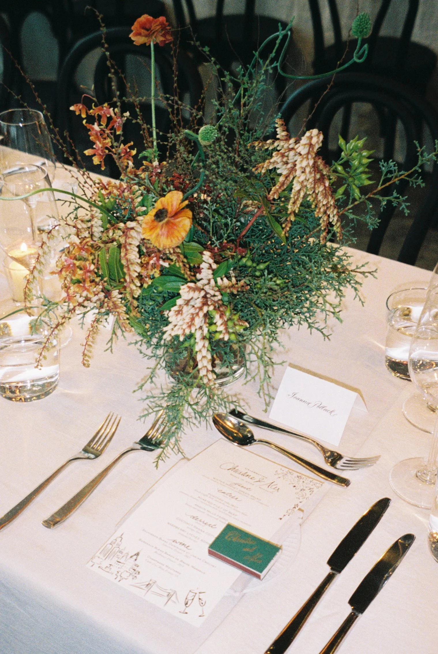 Elegant table setting with a floral centerpiece, silverware, glassware, and menu cards on a white tablecloth, with a black chair in the background.