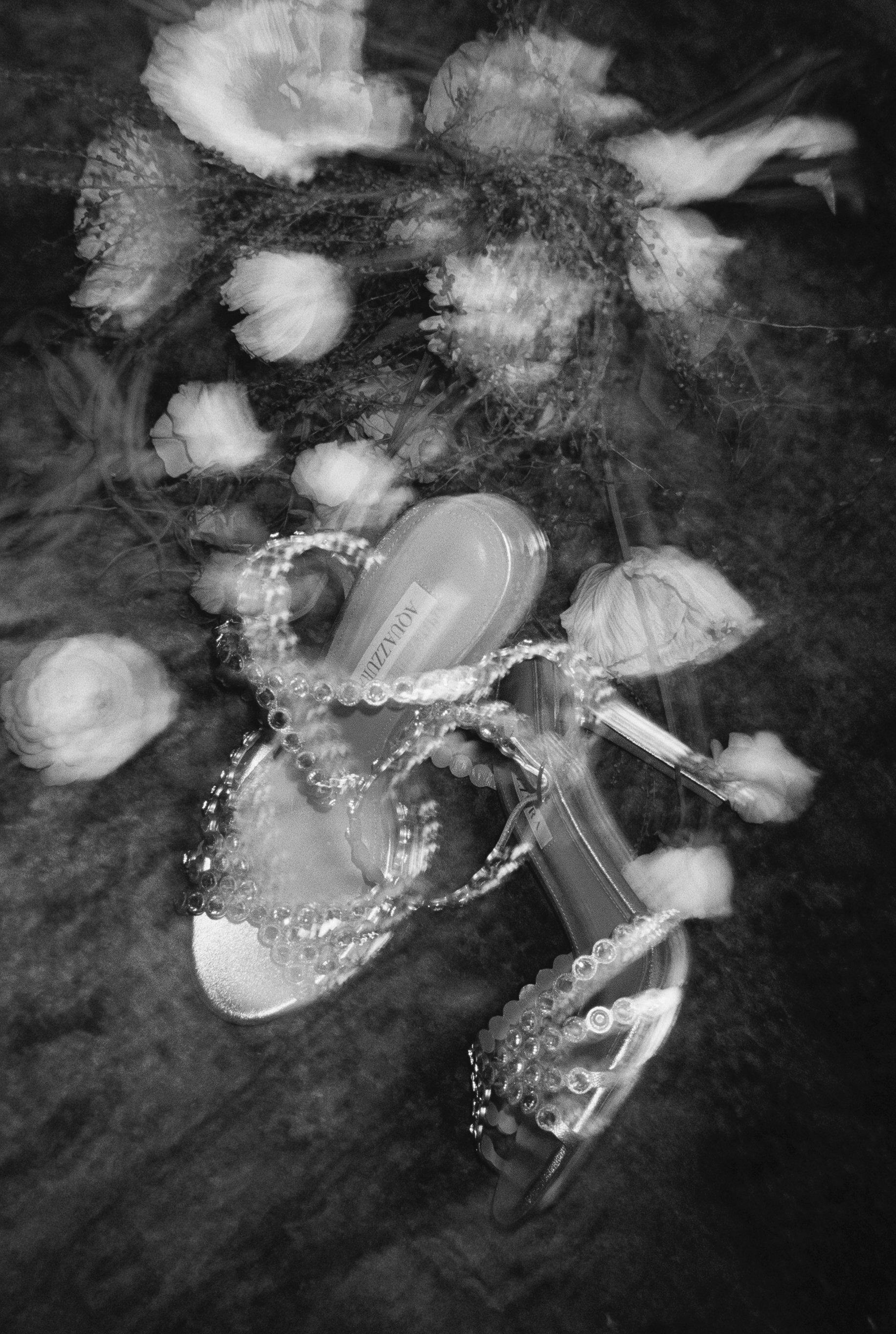 Black and white photo of flowers, a jewelry box with a chain, and a small container with a label, all placed on a textured surface.