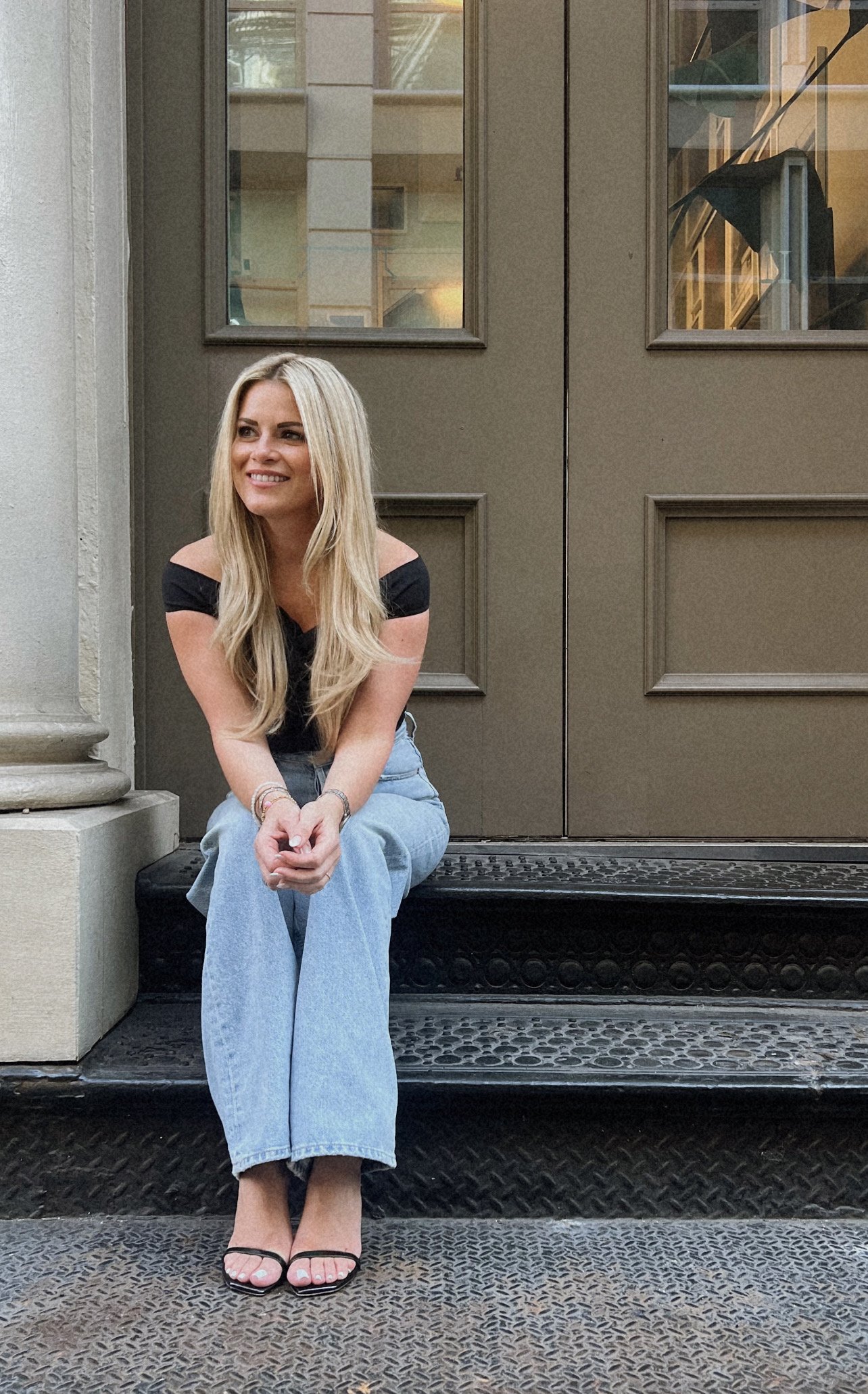 A woman with long blonde hair sitting on a step in front of a large brown door with windows, smiling and looking to her left, wearing a black off-shoulder top, light blue jeans, and black strappy sandals.