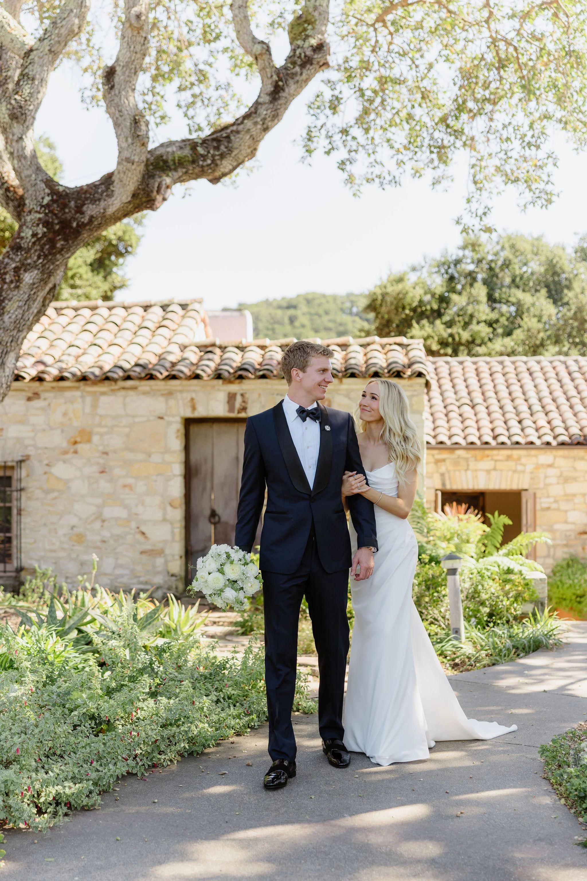 A bride and groom walk arm in arm outdoors in front of a stone building with a tiled roof, surrounded by green plants, trees, and flowers, with sunlight filtering through the leaves.