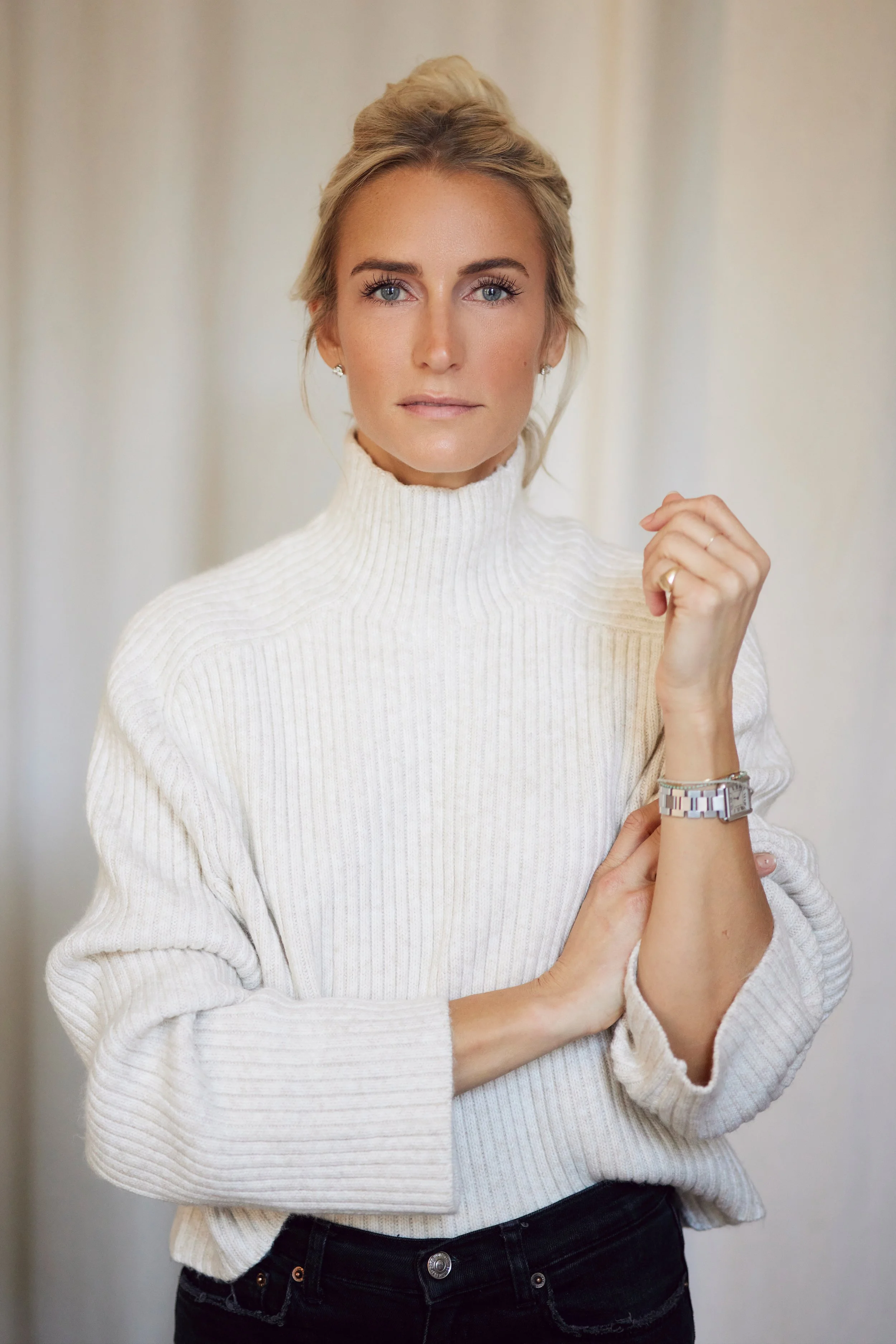 A woman with blonde hair tied up, wearing a white turtleneck sweater, posing with one hand near her face, wearing a watch and rings, against a plain background.