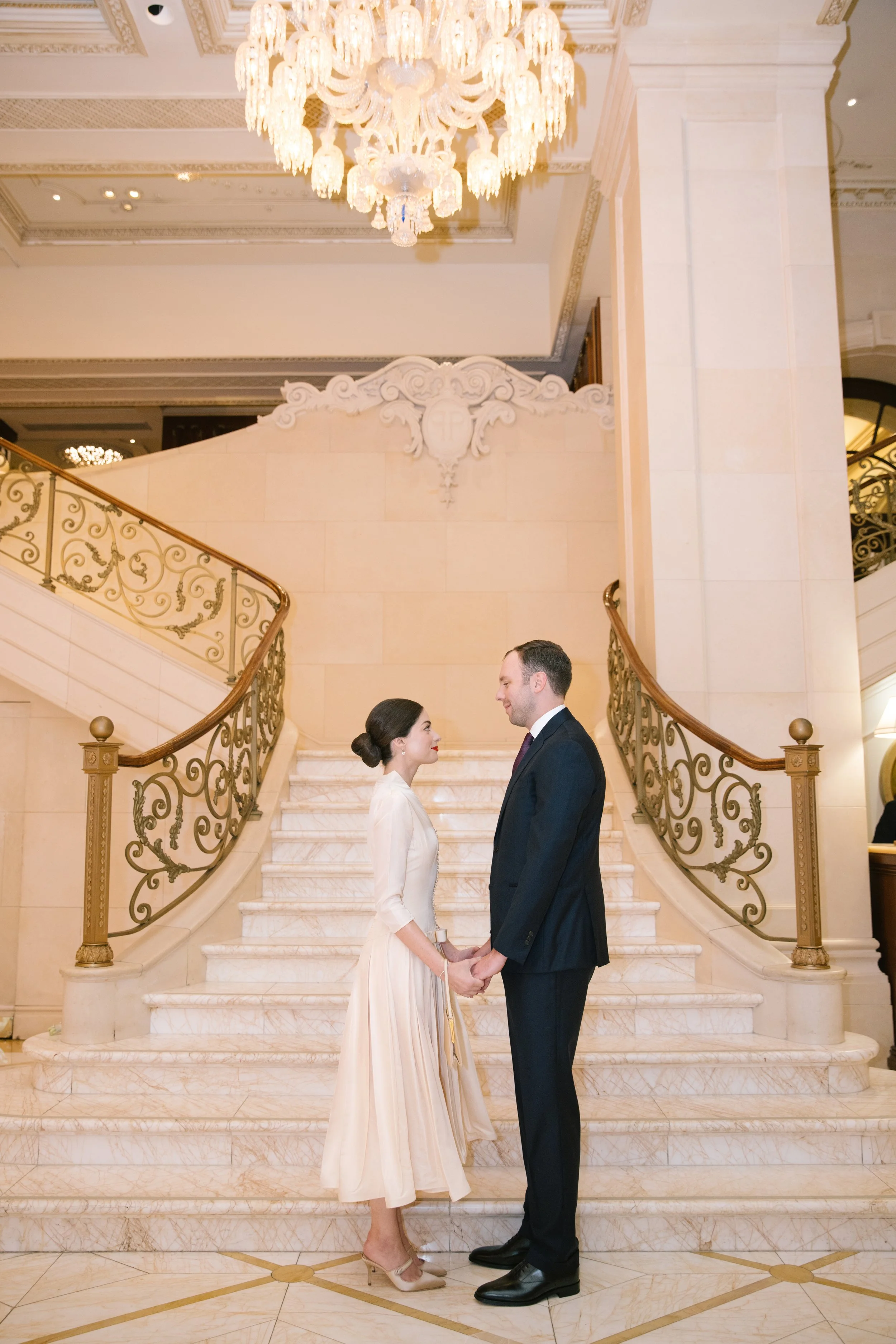 A couple dressed in wedding attire stand face to face holding hands on a marble staircase in an elegant, ornate ballroom with a large crystal chandelier overhead.