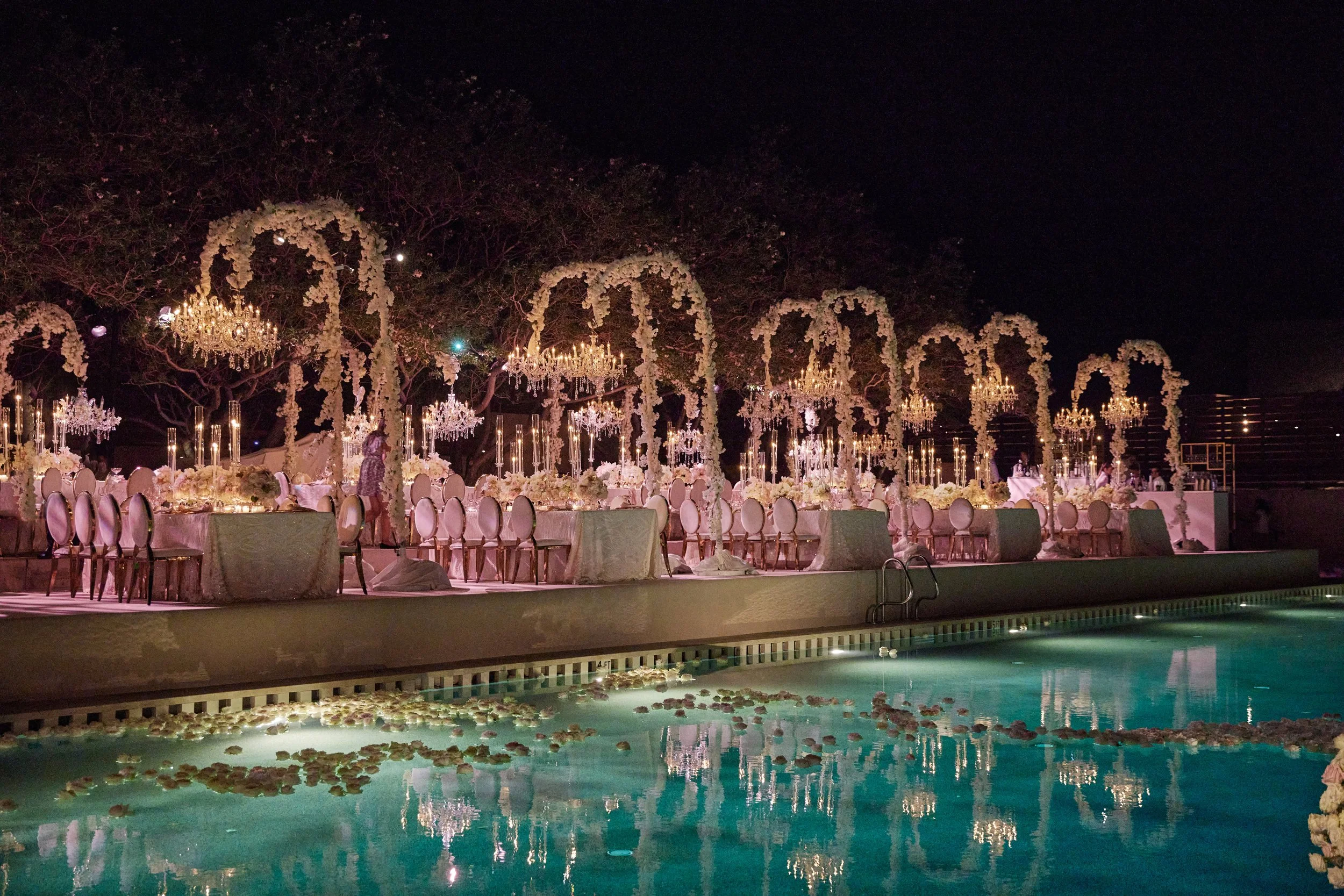 Nighttime wedding reception setup by a pool with decorated tables, chandeliers, floral arches, and hanging candles surrounded by cast iron chairs.