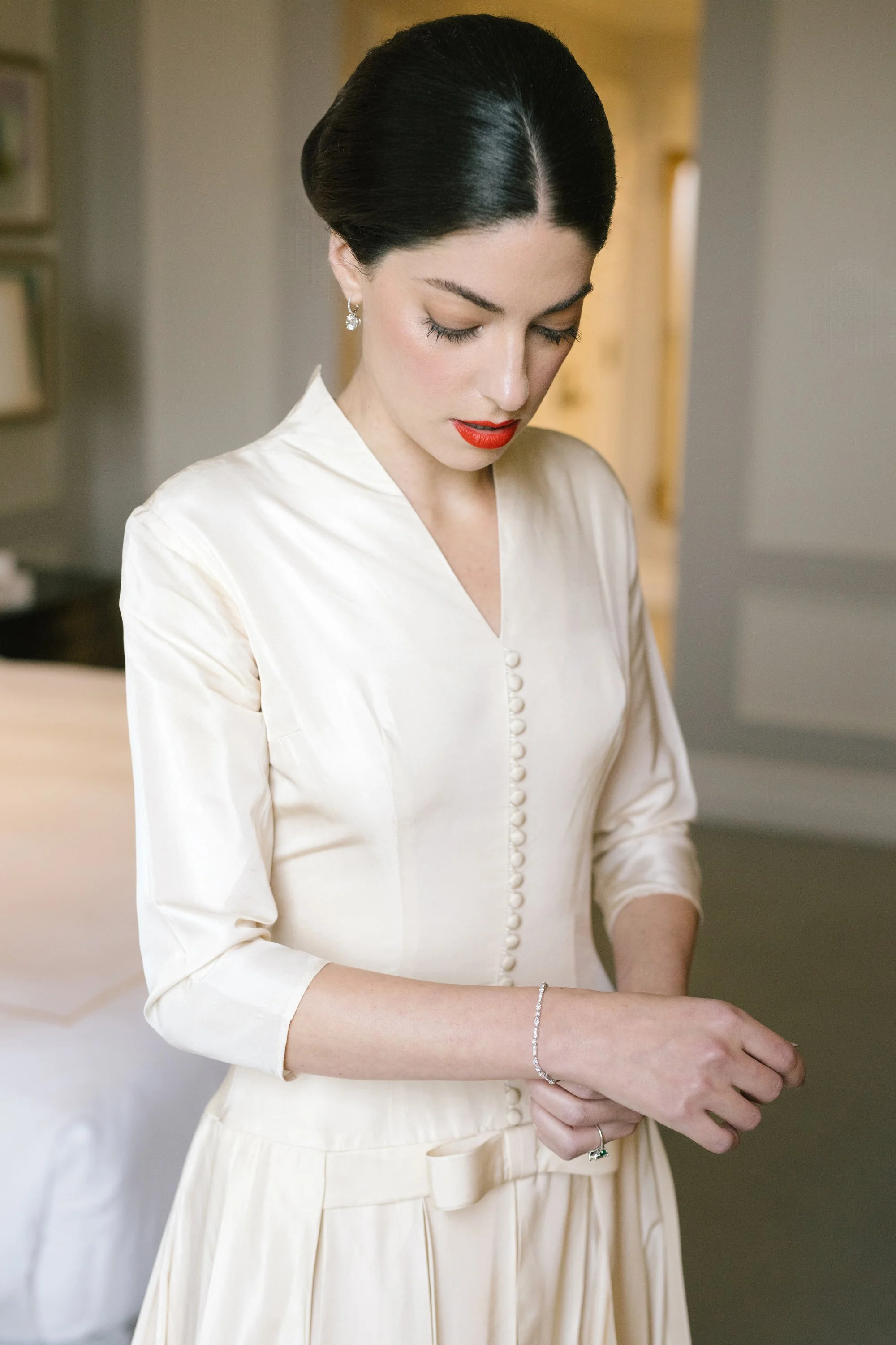 A woman with dark hair styled in an elegant updo wearing a cream-colored vintage dress, red lipstick, earrings, and a bracelet, standing indoors with her eyes closed and hands clasped.