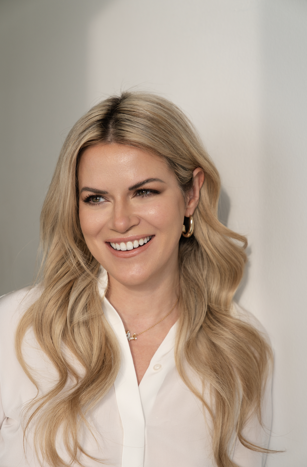 A woman with long blonde wavy hair, smiling and wearing a white blouse, gold hoop earrings, and a delicate necklace, standing against a plain light-colored wall.