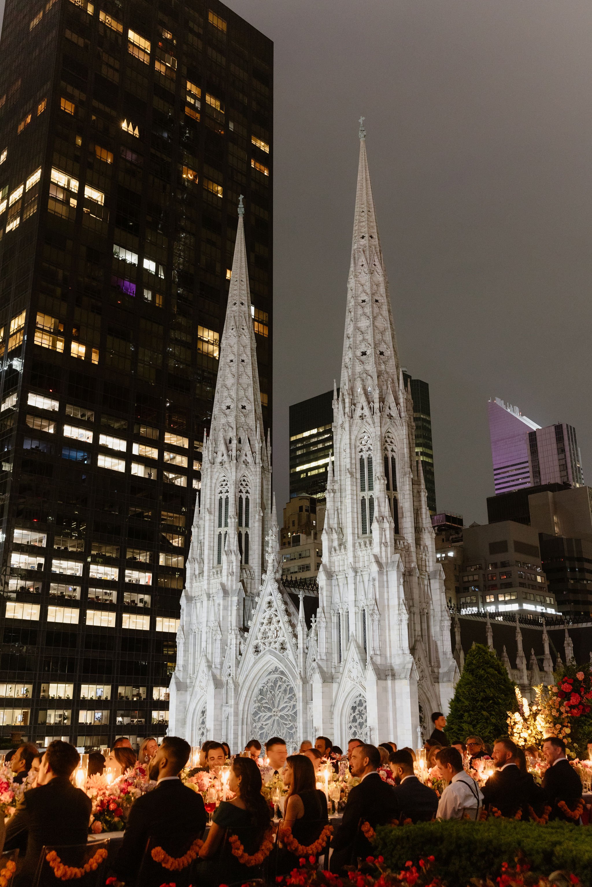 The image shows a nighttime scene of a Gothic-style church with tall spires, situated among modern high-rise buildings. In front of the church, there is a large outdoor gathering of people dressed formally, sitting at long tables decorated with flowe