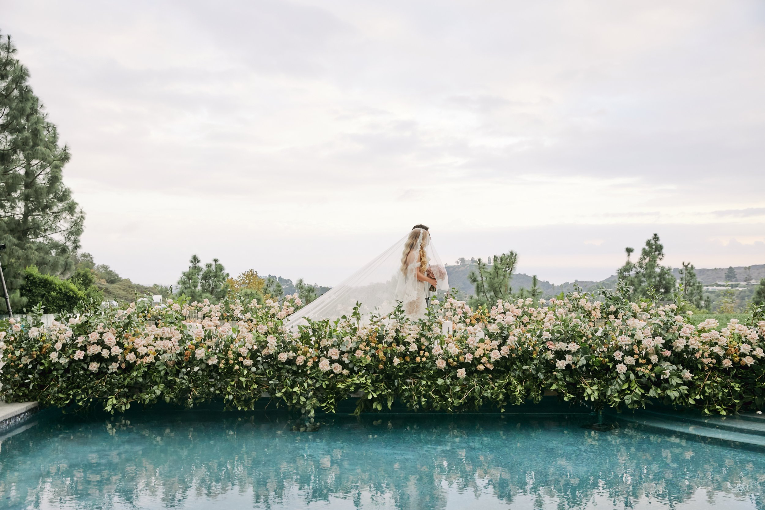 A bride holding a bouquet stands in a garden filled with pink roses, with a water feature in the foreground and trees and cloudy sky in the background.