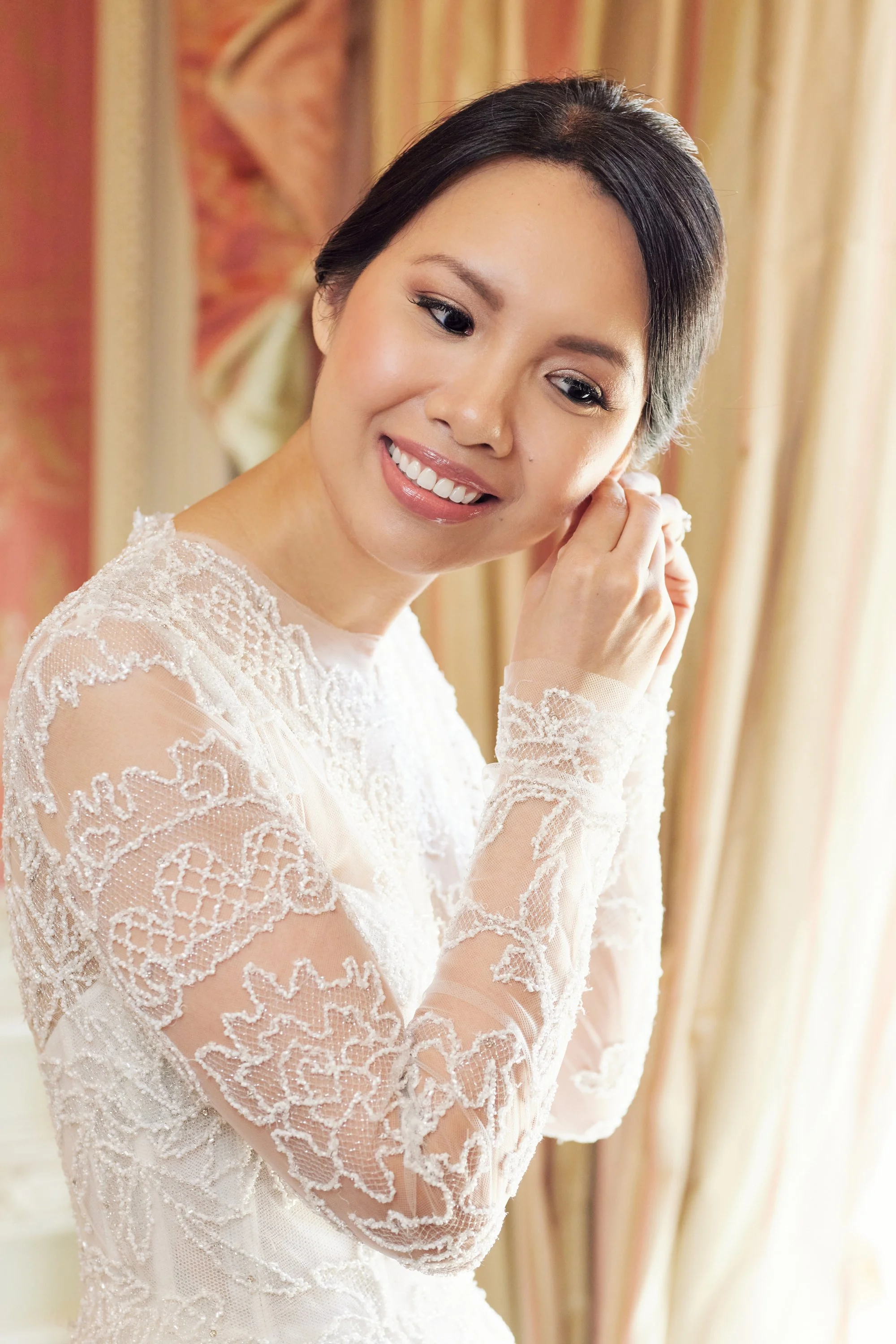 A woman with short black hair is smiling while putting on an earring. She is wearing a white lace dress with intricate embroidery and sheer long sleeves. The background features warm-colored curtains.