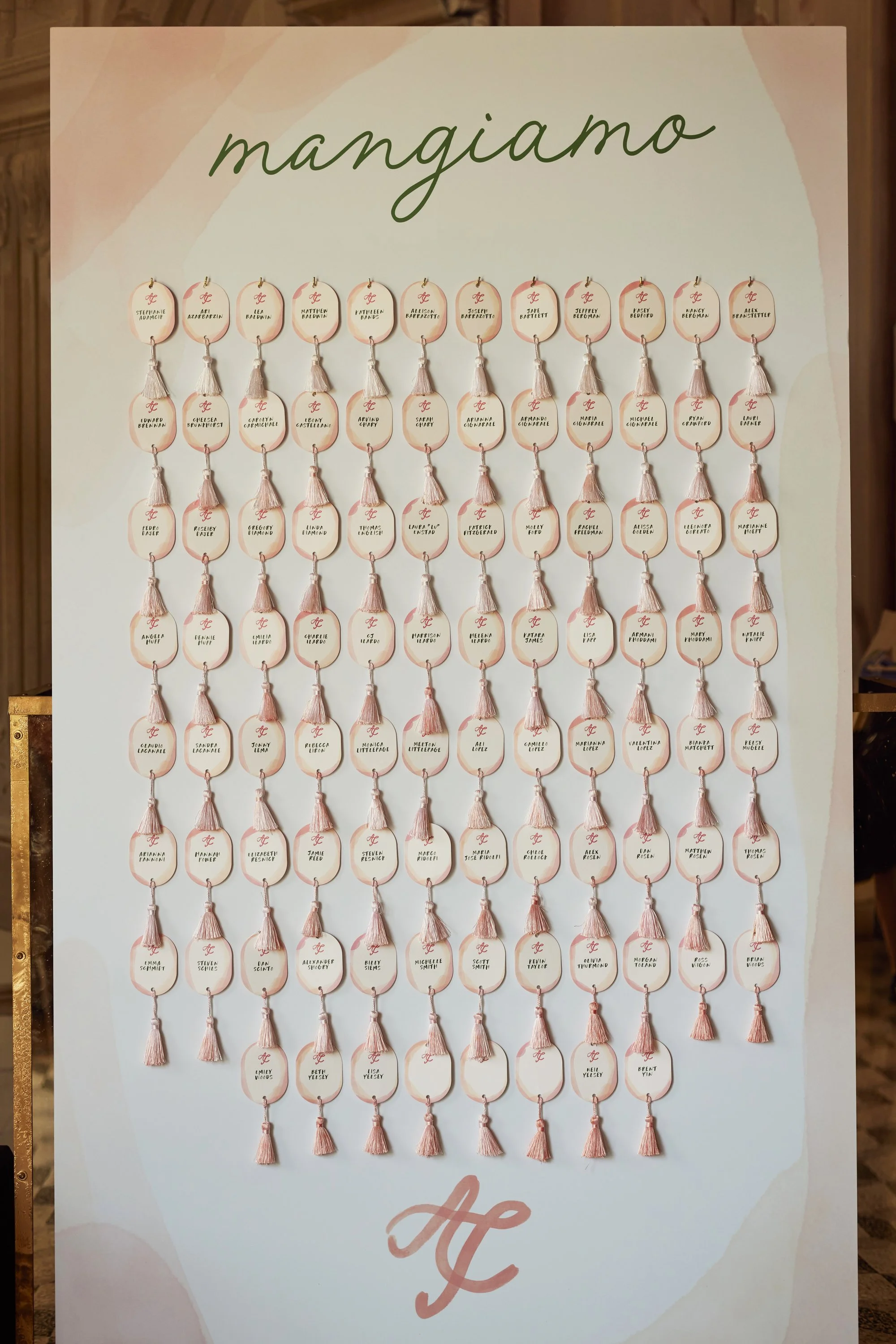 A large display board with multiple rows of small oval tags, each featuring a name and a small pink tassel. The top of the display has the word 'mANGiamo' written in cursive, with the bottom corner showing a stylized letter 'A' and the word 'love'.