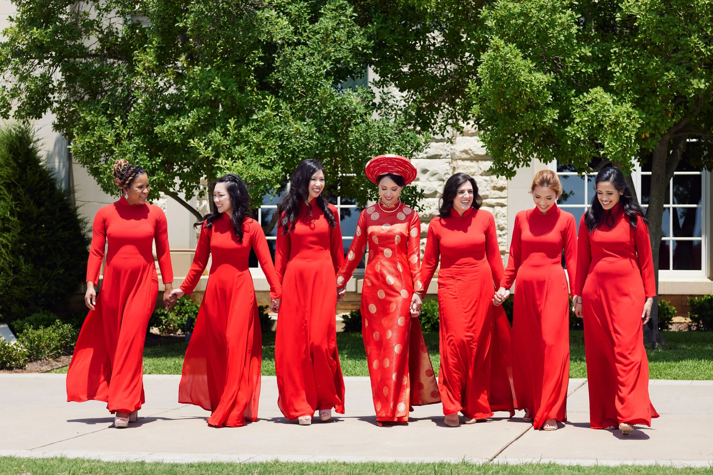 A group of seven women in red dresses walking hand in hand outdoors, with trees and a building in the background.