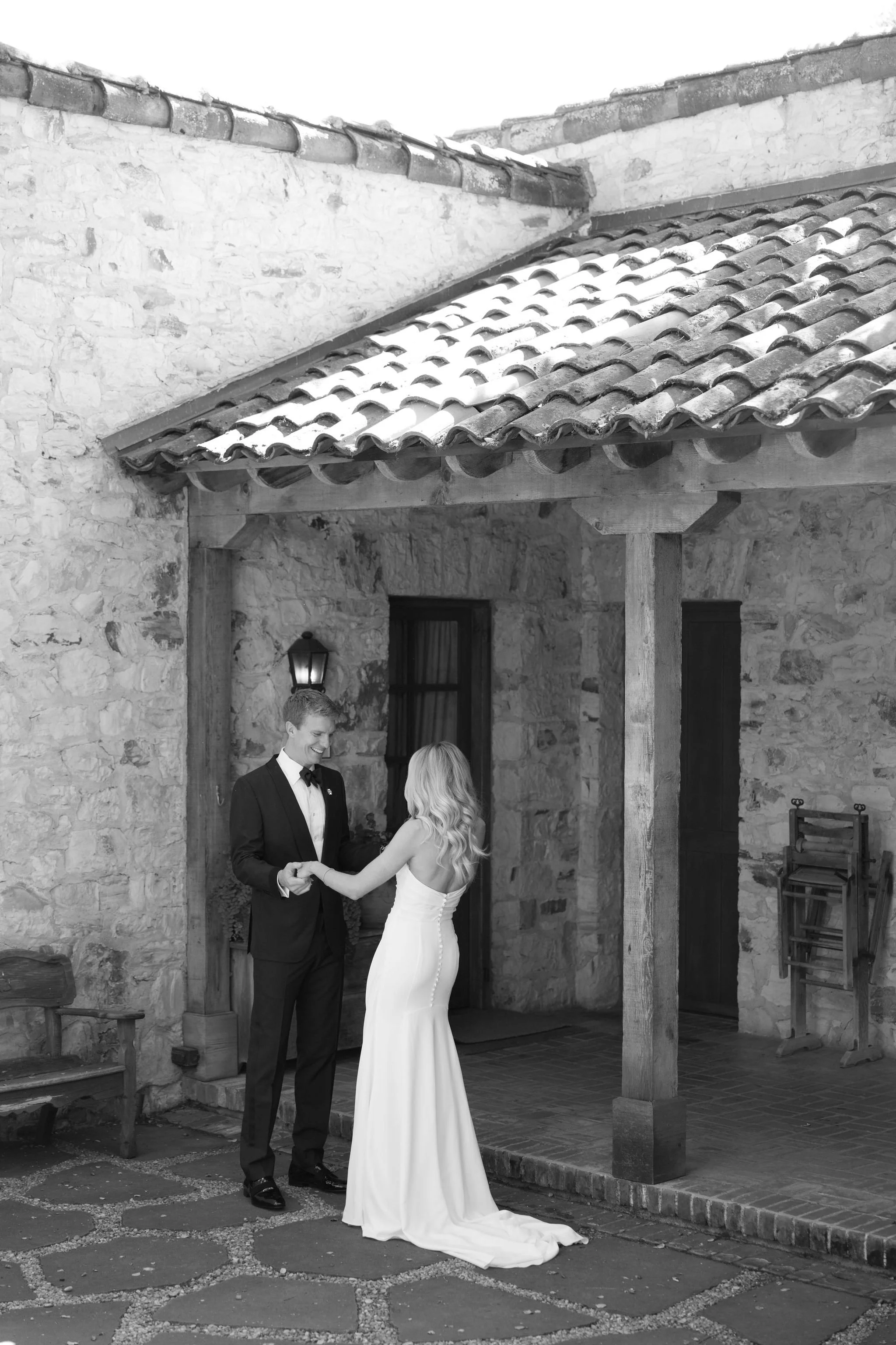 A couple exchanging vows during a wedding ceremony outdoors by a rustic stone building, with the groom in a tuxedo and the bride in a white wedding dress, holding hands.