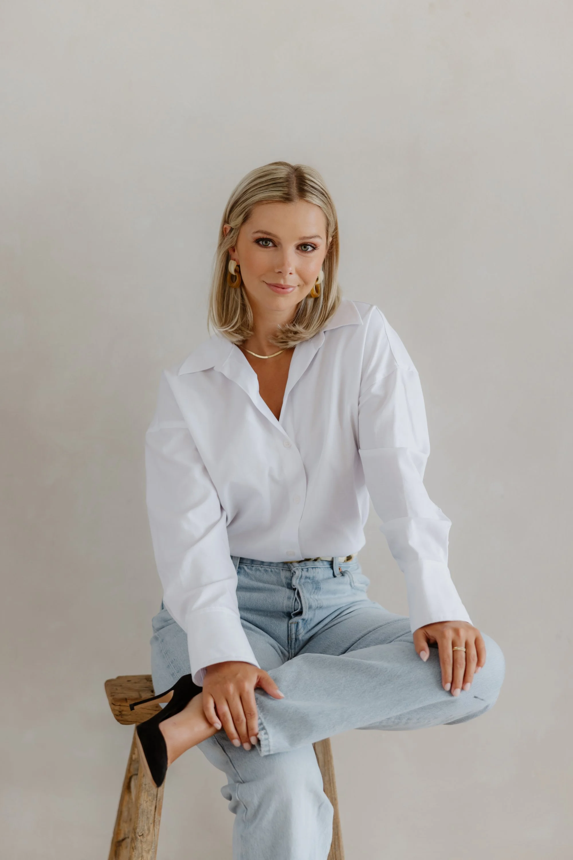 A woman with shoulder-length blonde hair, wearing a white button-up shirt, light blue jeans, and black high heels, sitting cross-legged on a wooden stool against a plain beige background, looking at the camera with a slight smile.