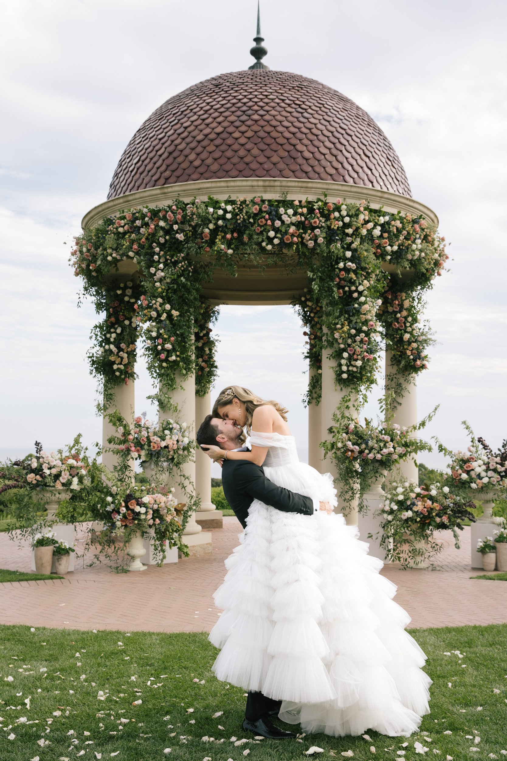 A bride and groom are sharing a kiss and dance beneath a floral-draped gazebo with a domed roof, columns, and floral arrangements, during a wedding celebration outside.