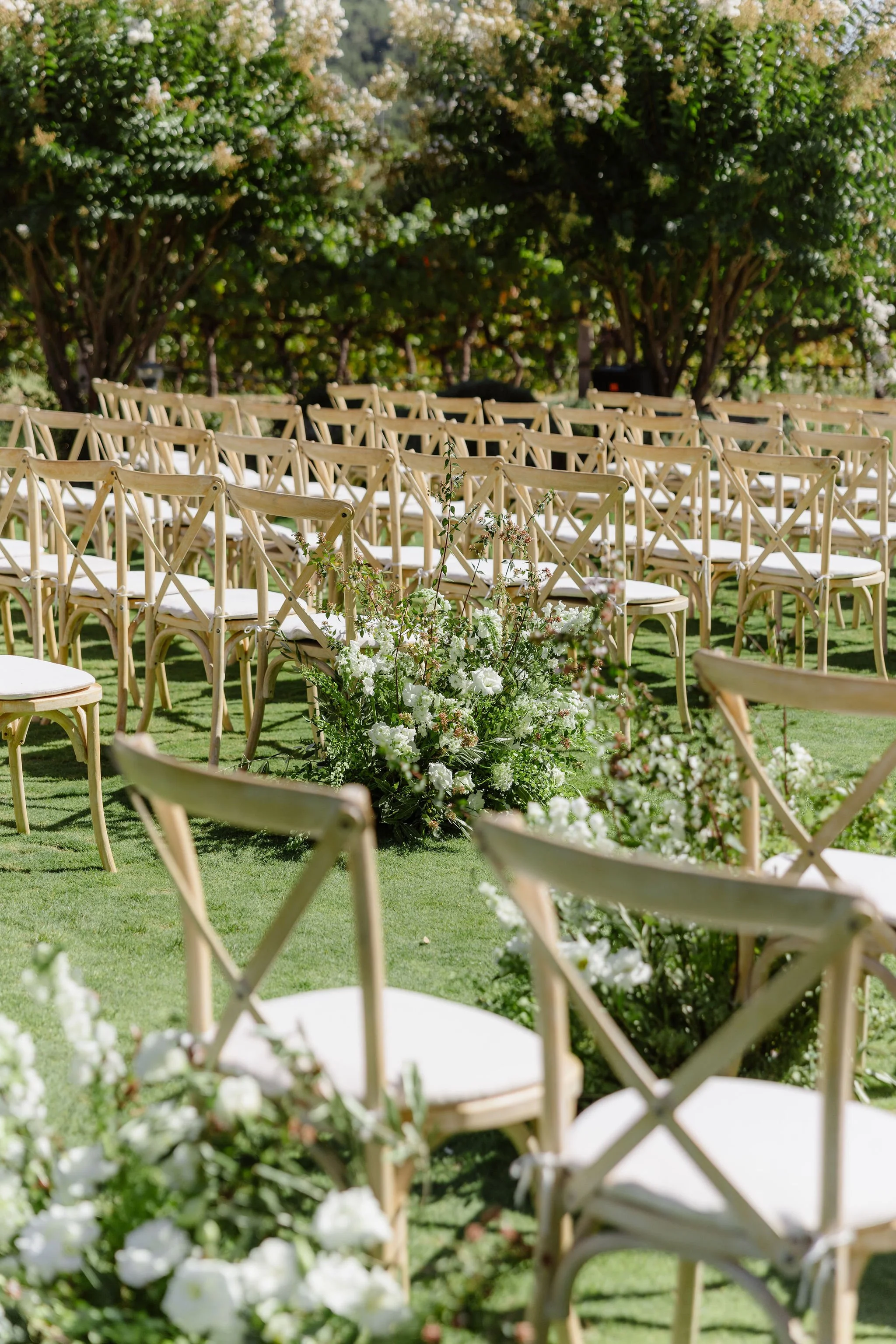 Outdoor wedding ceremony setup with rows of wooden chairs and floral arrangements under trees.