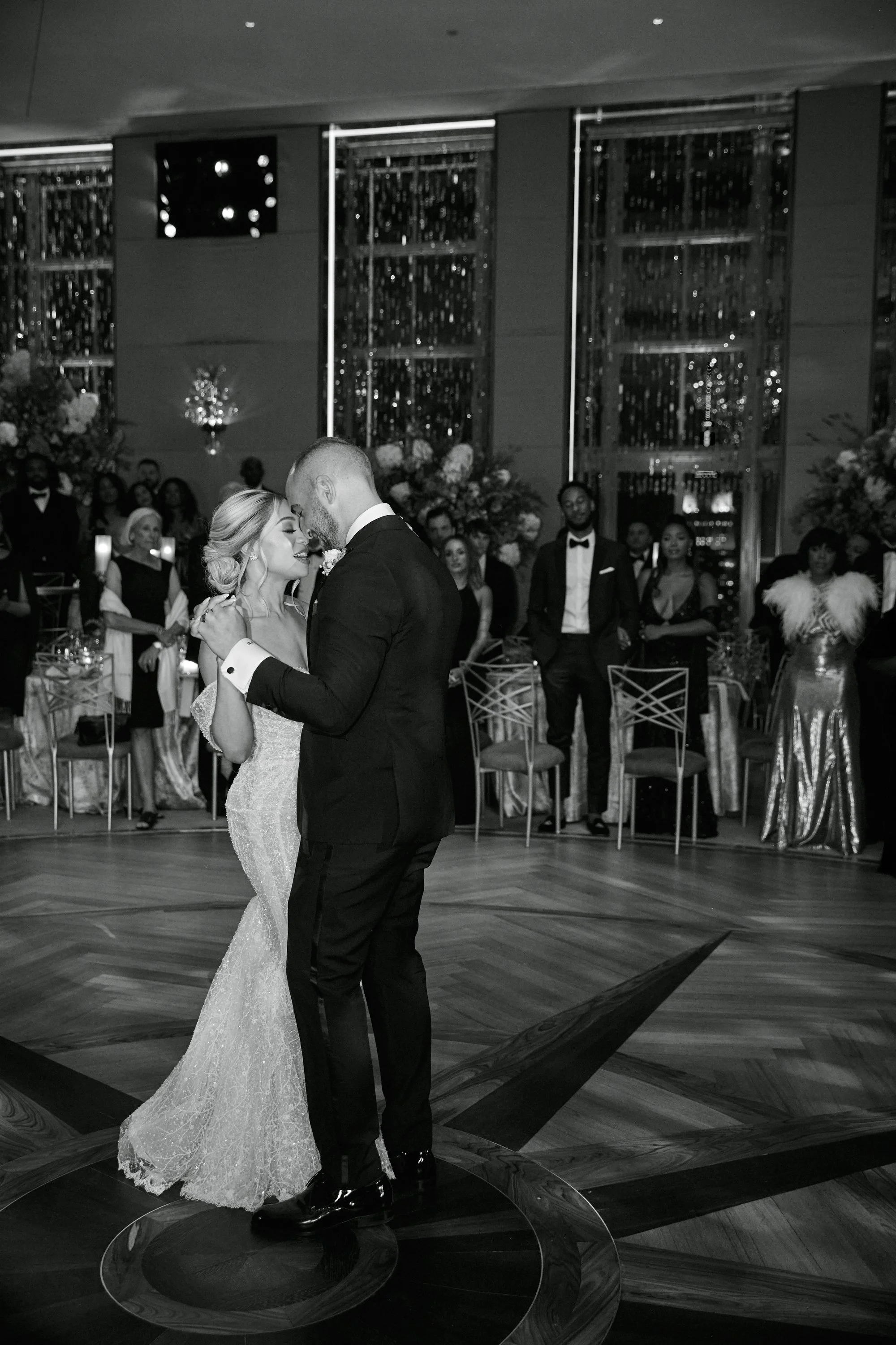 A bride and groom share a dance at their wedding reception with guests watching in the background. The black and white photo captures a joyful moment.