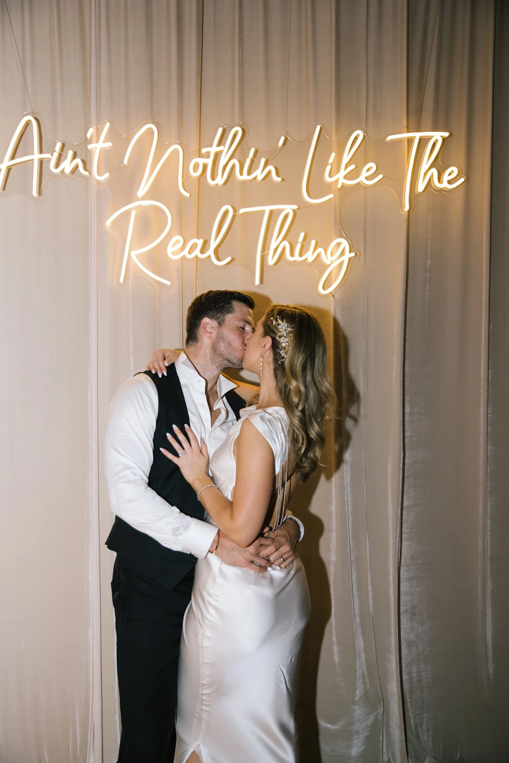 A couple dressed in wedding attire kissing in front of a beige curtain with a neon sign that reads "Ain't Nothin' Like The Real Thing."