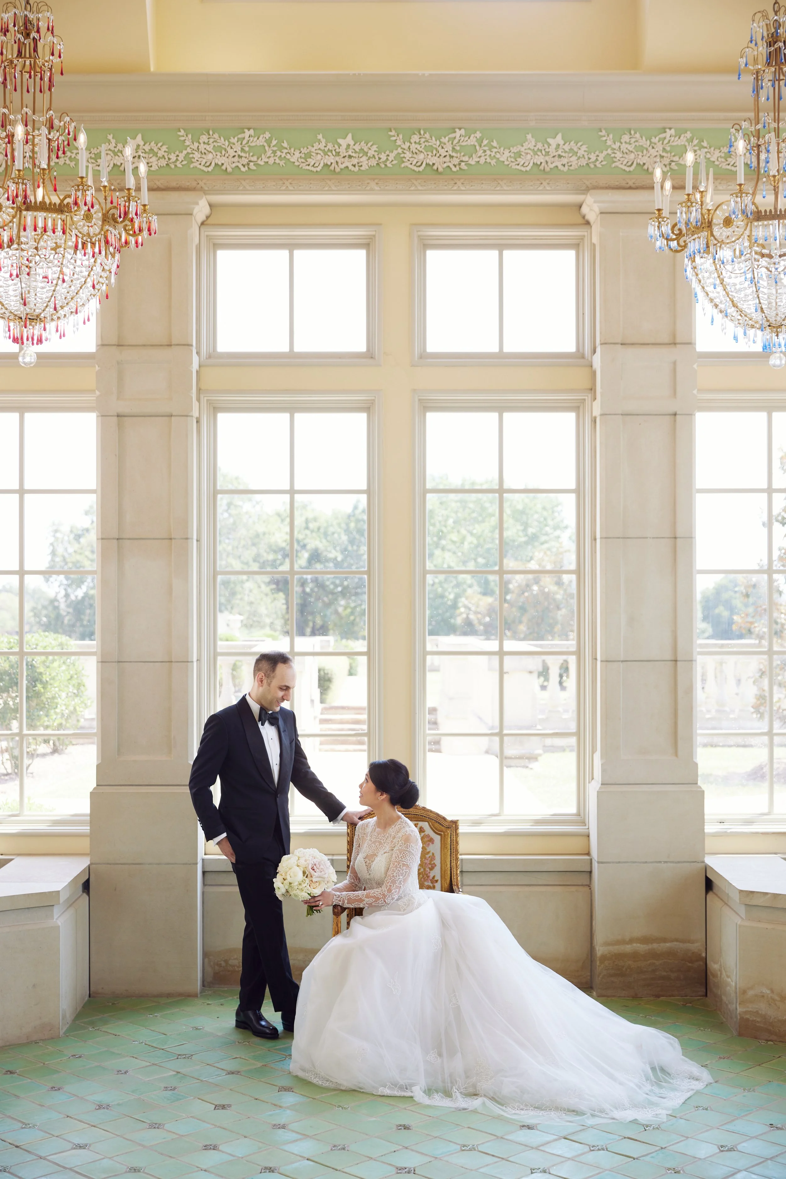 A wedding reception scene with a bride and groom indoors by large windows and chandeliers. The groom in a black tuxedo is standing next to the seated bride in a white wedding gown holding a bouquet of flowers. The bride is looking up at the groom as they share a moment.