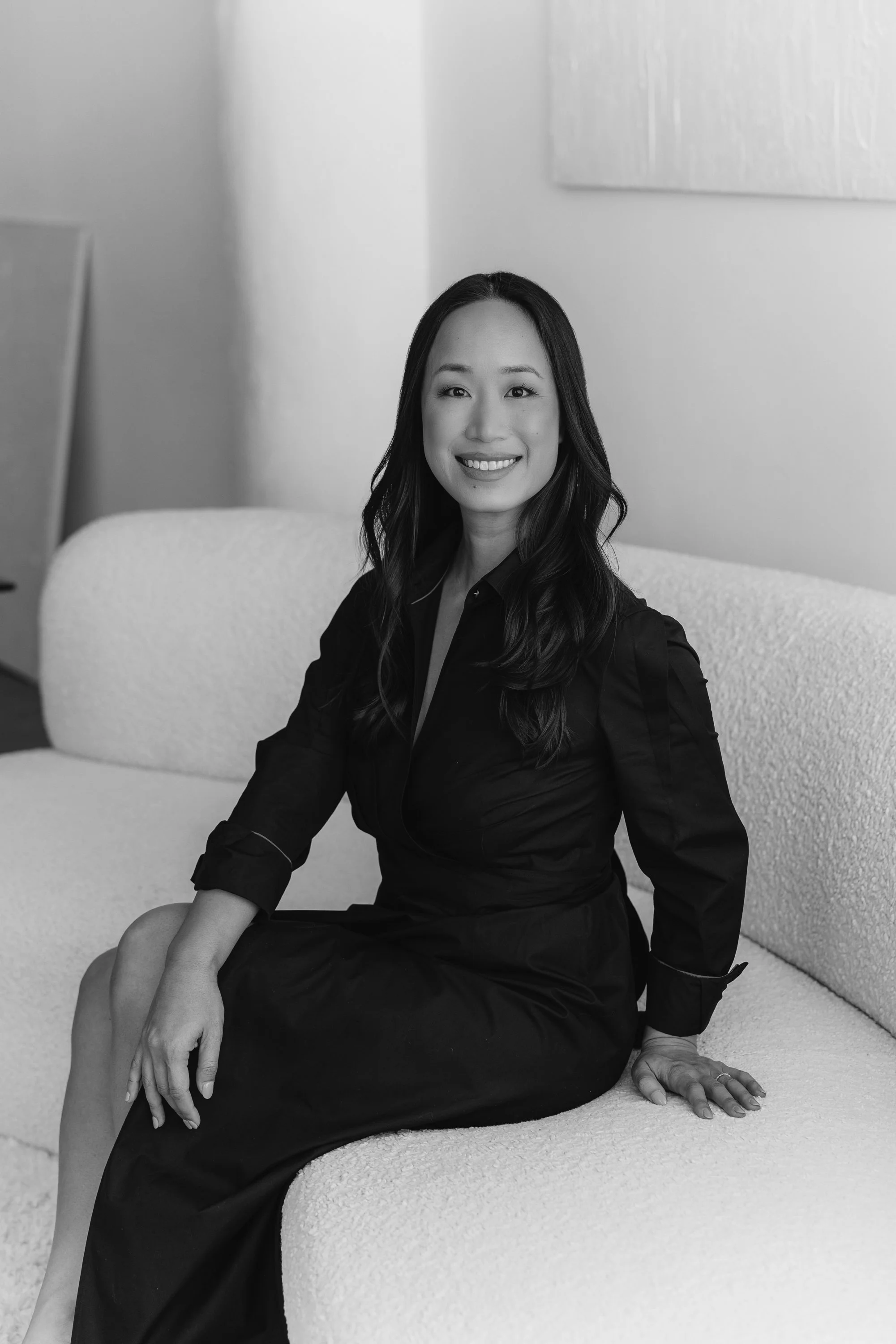 Black and white photo of a woman with long wavy hair, sitting on a textured sofa in a room with minimal decor, smiling at the camera. She is wearing a dark outfit with rolled-up sleeves.