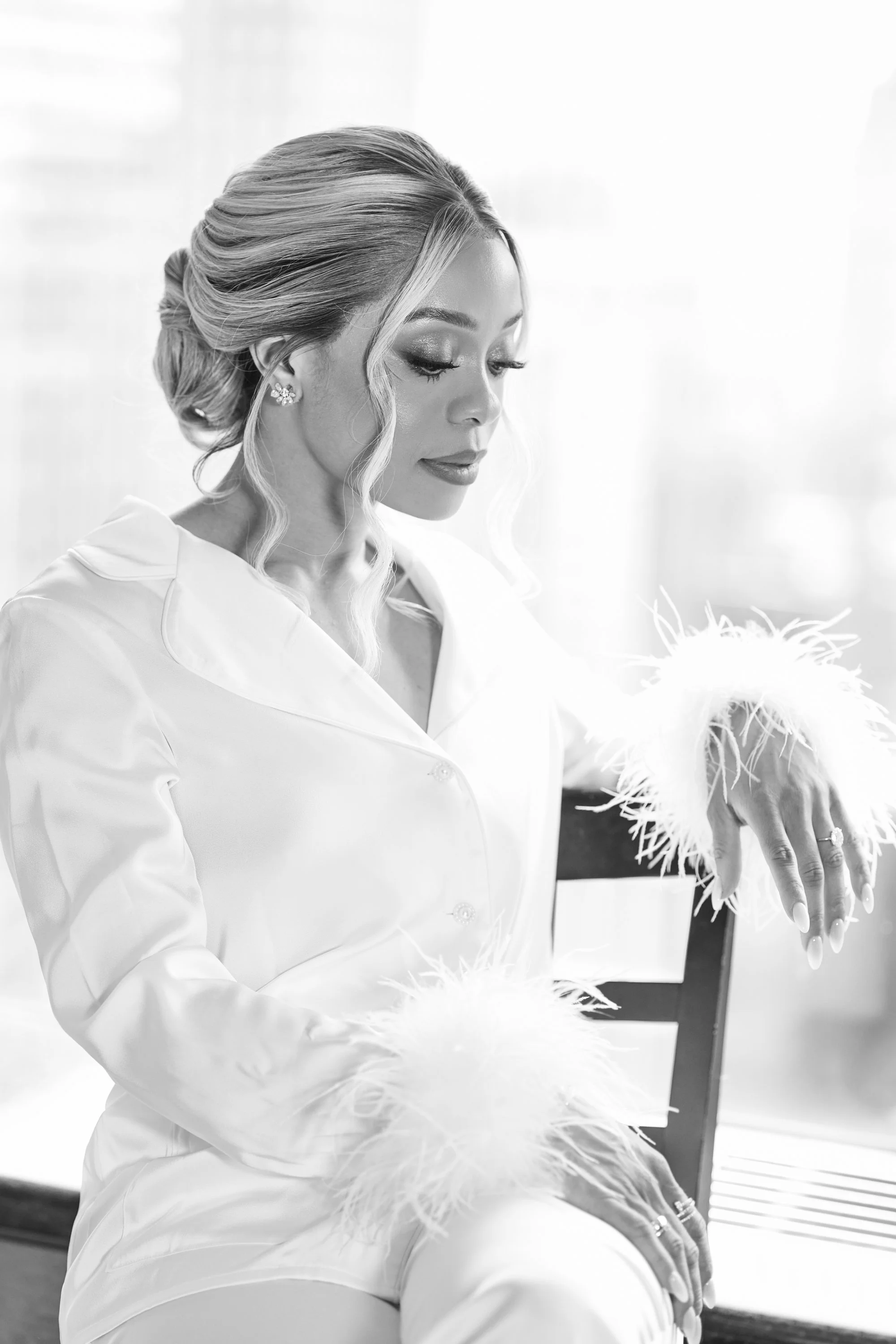 Black and white photo of a woman with styled hair and makeup, wearing a satin robe with feathered cuffs, sitting on a chair near a window with blinds.