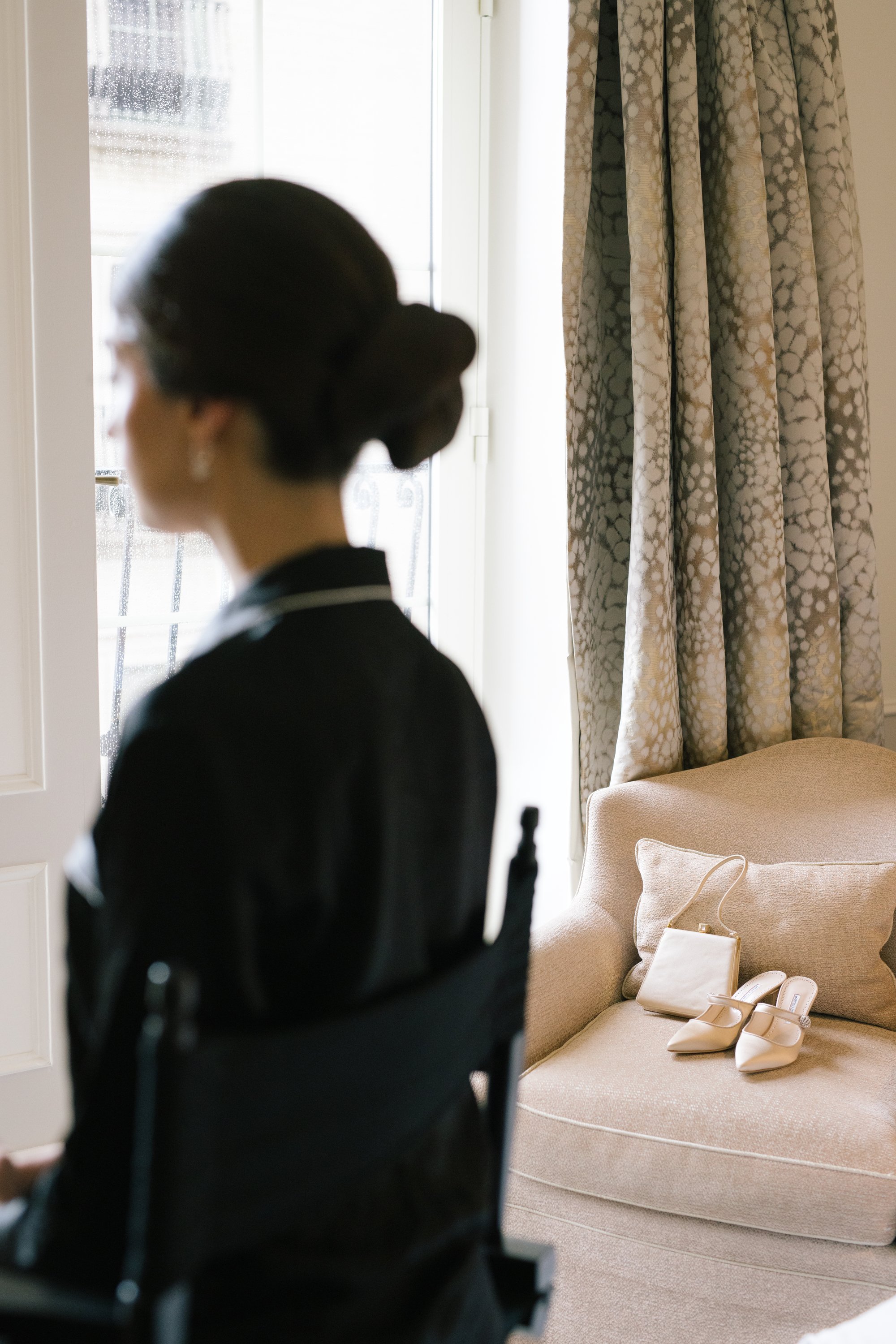 A woman with dark hair styled in an elegant updo, wearing earrings and a dark blazer with white trim, is standing near a doorway inside a room. In the background, a beige sofa on which a pair of white high-heeled shoes, a bag, and a phone are placed can be seen. The room has patterned curtains and bright natural light coming through the window.
