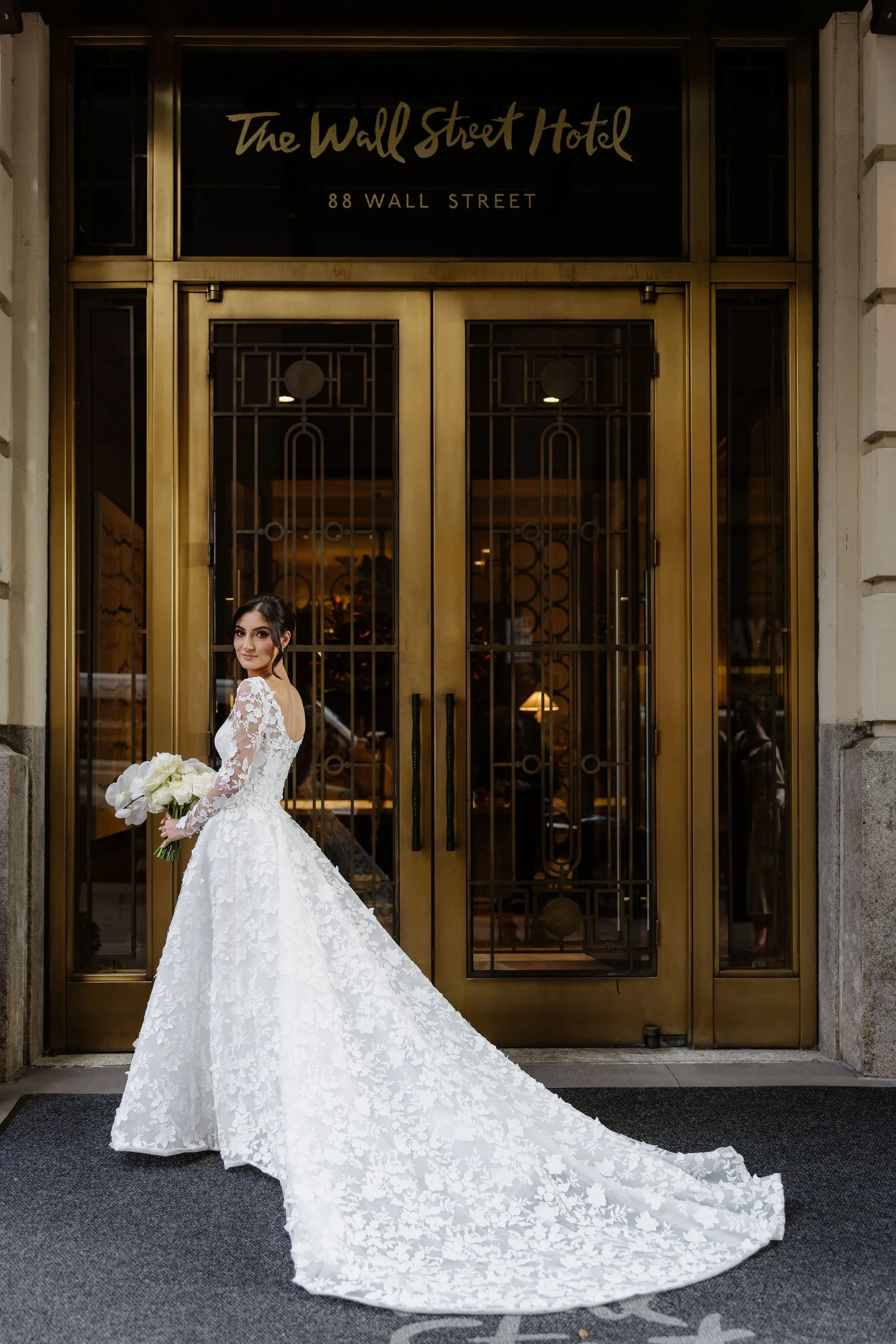 A bride in a white lace wedding gown holding a bouquet of white flowers, standing in front of a gold-colored double door entrance to The Wall Street Hotel at 88 Wall Street.