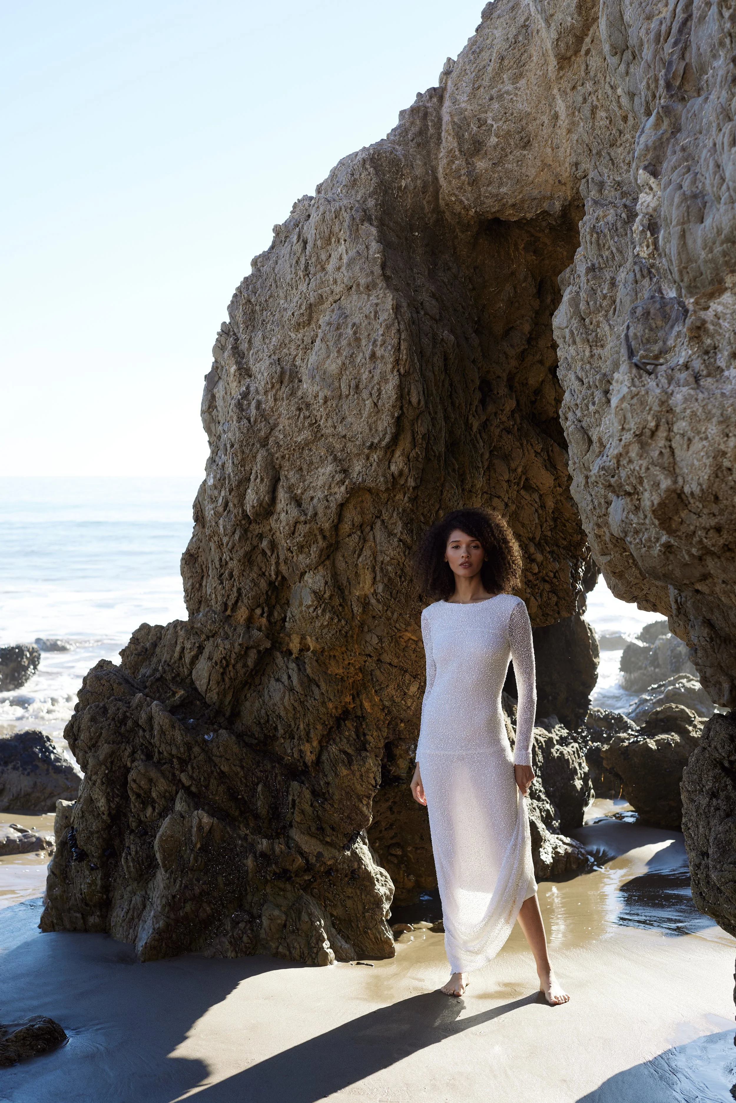 Woman in a long white dress standing on a sandy beach near large rock formations with the ocean and clear sky in the background.