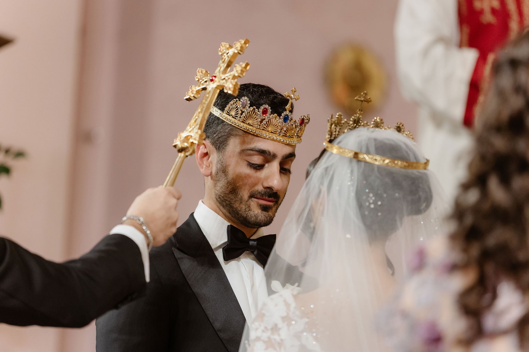 A groom in a black tuxedo and bow tie is crowning a bride with a gold crown during their wedding ceremony.