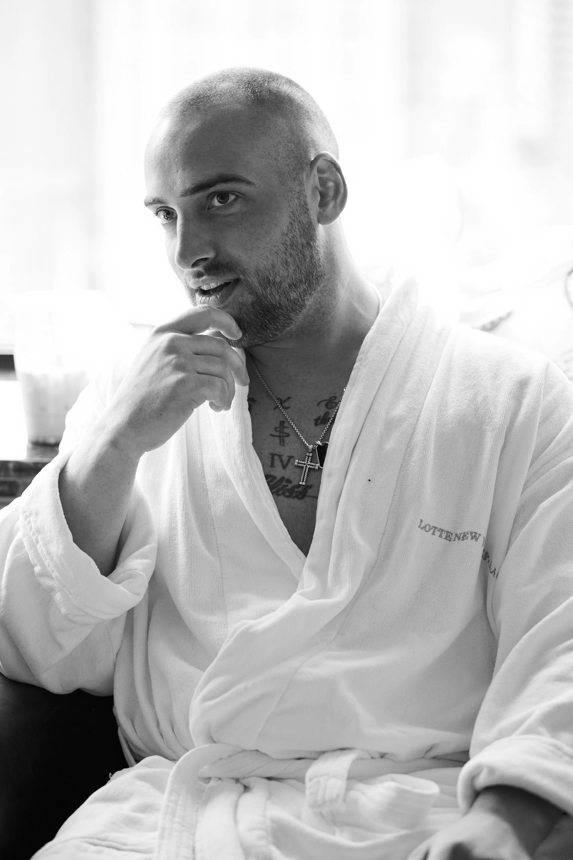 Black and white photo of a man with a shaved head and beard wearing a martial arts gi, sitting with one hand near his mouth, with visible tattoos on his chest and neck, and a cross necklace. He appears to be deep in thought.