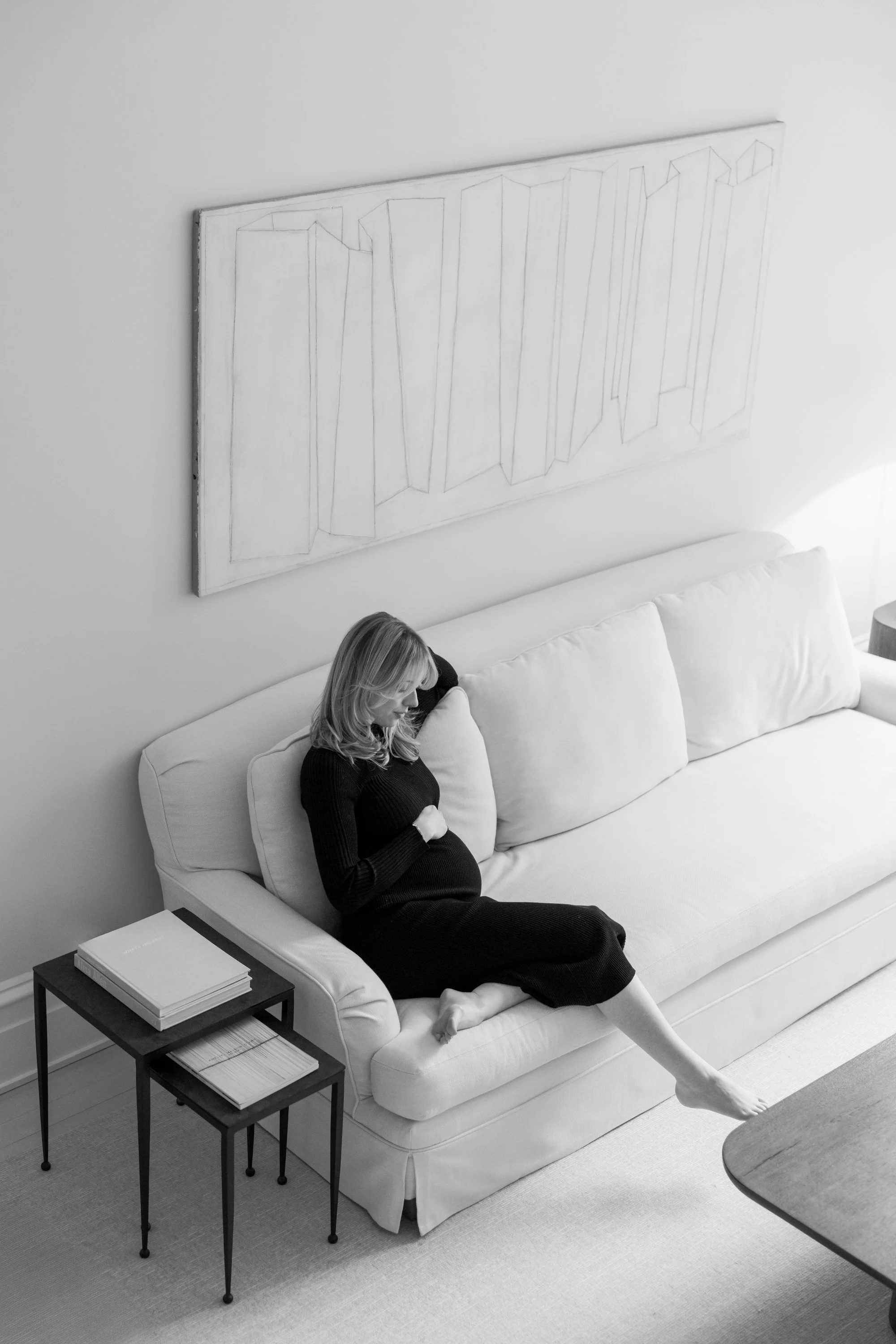A pregnant woman in a black dress resting on a white sofa in a minimalist living room, with a side table holding books and a stack of magazines, and abstract geometric artwork hanging on the wall.