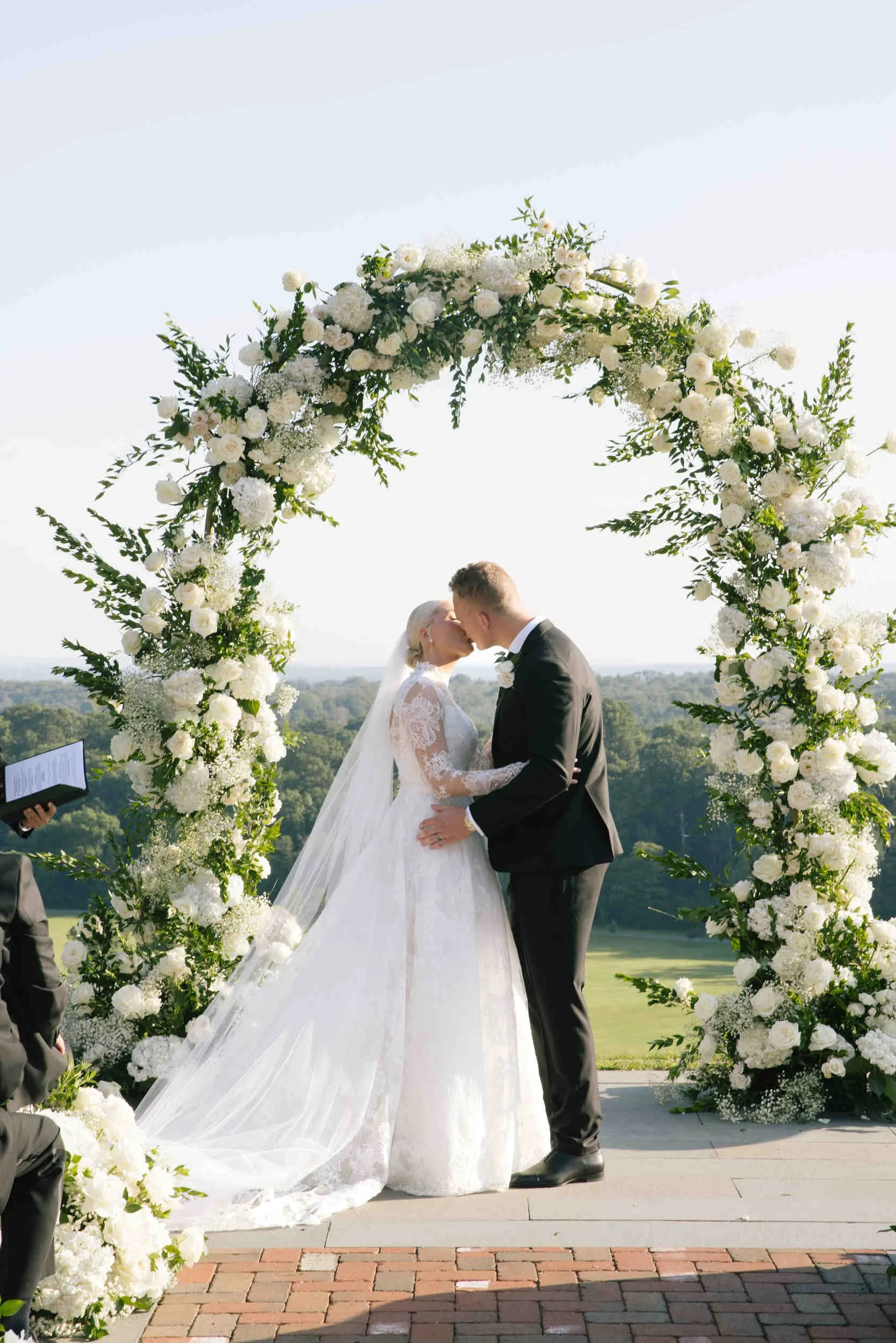 A couple in wedding attire kissing under a large floral arch at an outdoor wedding ceremony during daylight.